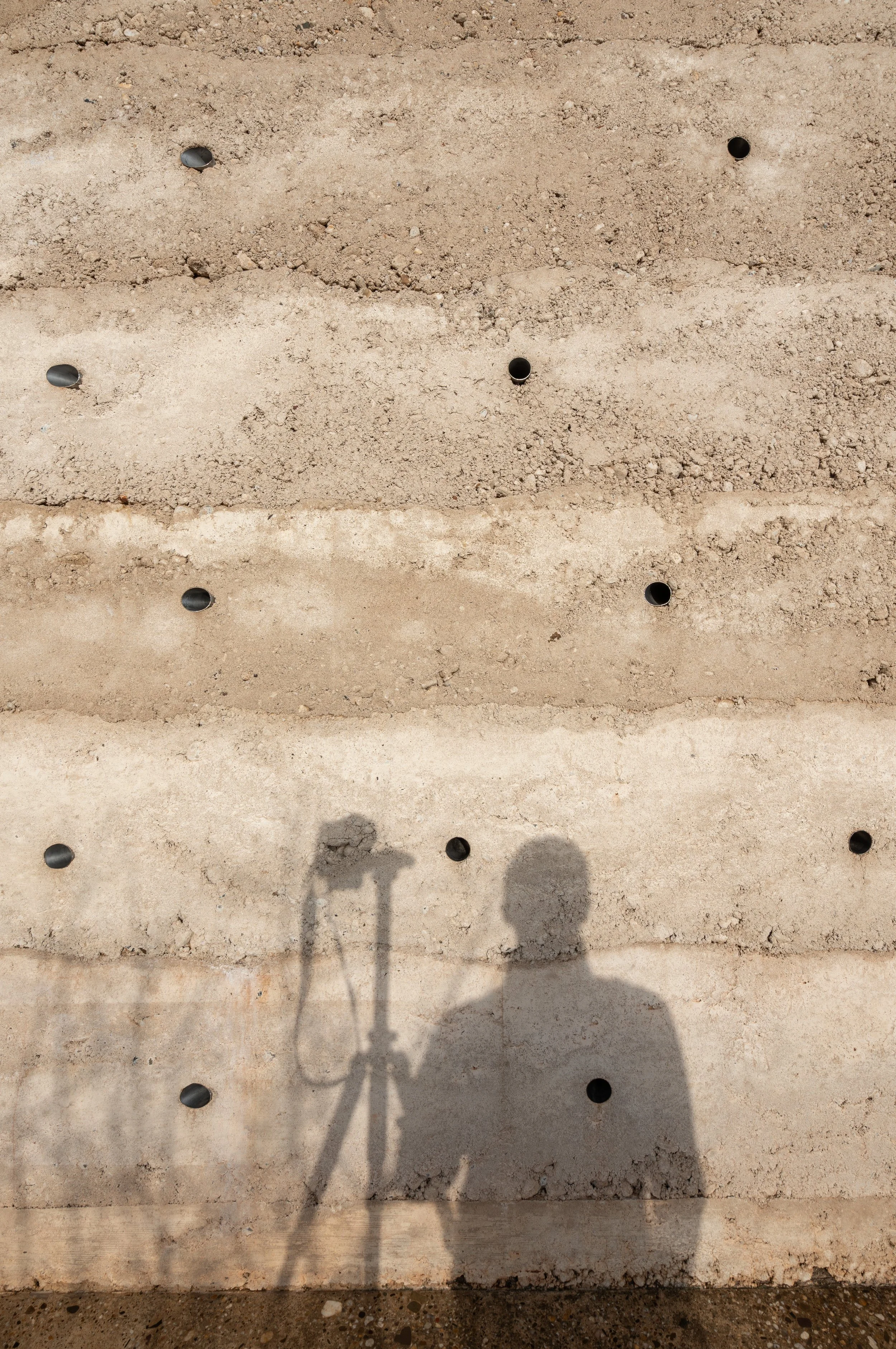 Der Schatten einer Person hält eine Kamera und steht vor einer Wand mit mehreren Reihenlochungen, die auf eine Baustelle hinweisen.