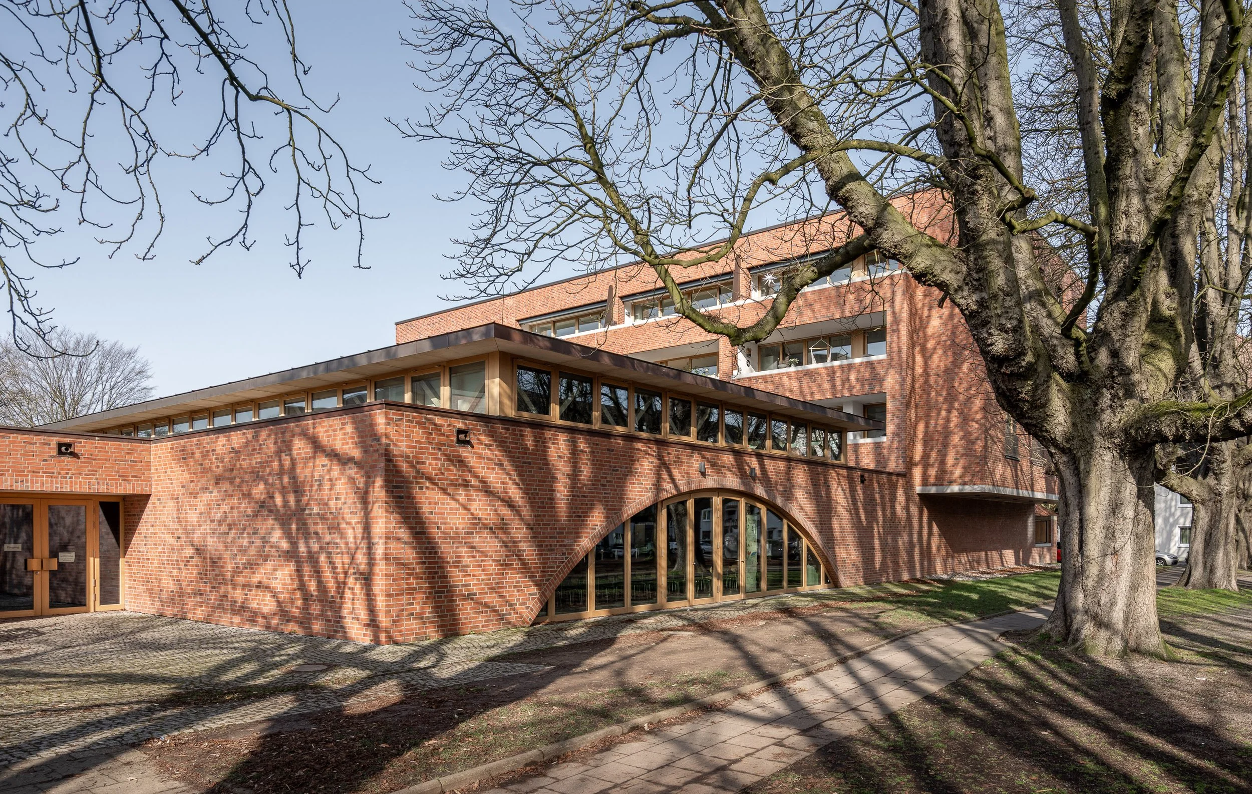 Moderne, mehrstöckige roten Backstein-Gebäude mit großen Fenstern und Bogen, umgeben von großen Bäumen im Sonnenschein.