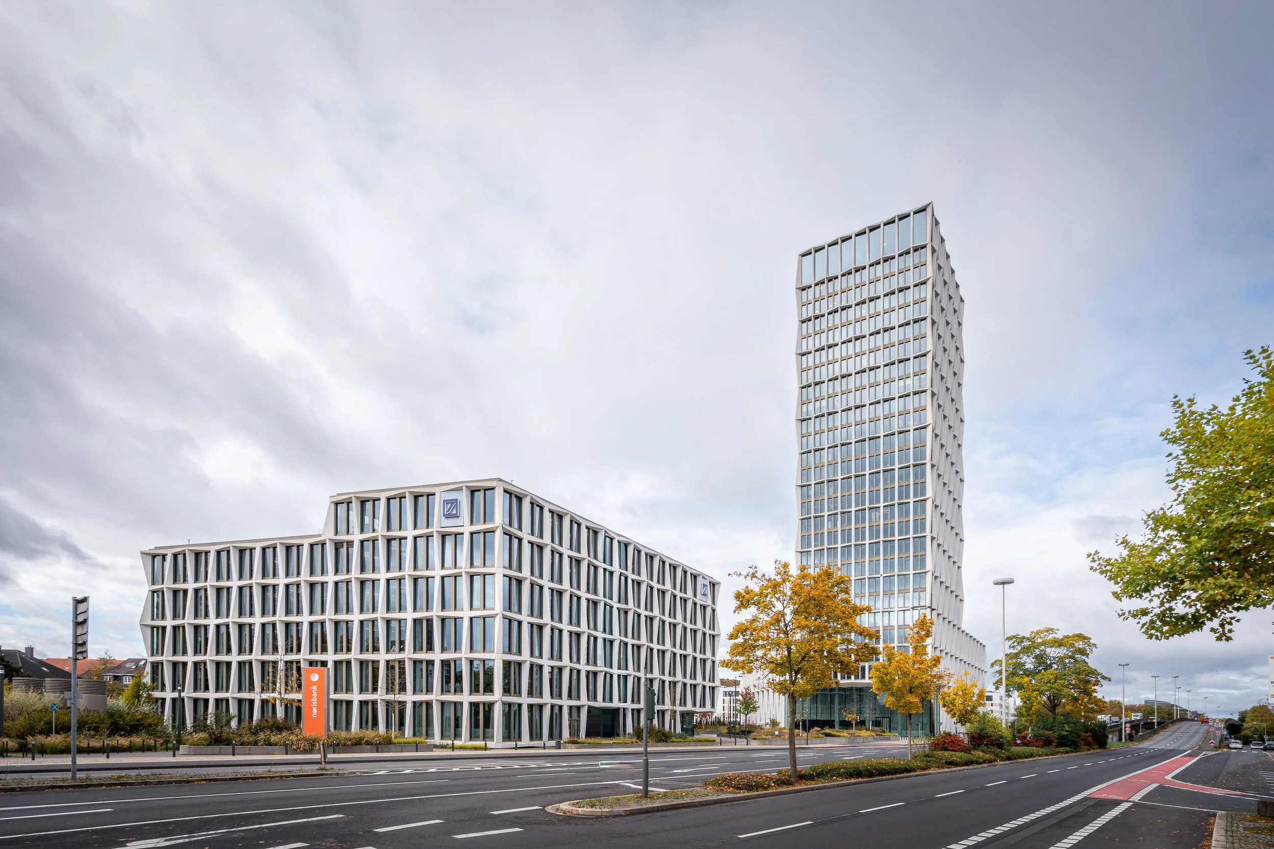 Moderne Bürogebäude neben einer Straße mit Bäumen im Herbst, bei bewölktem Himmel.