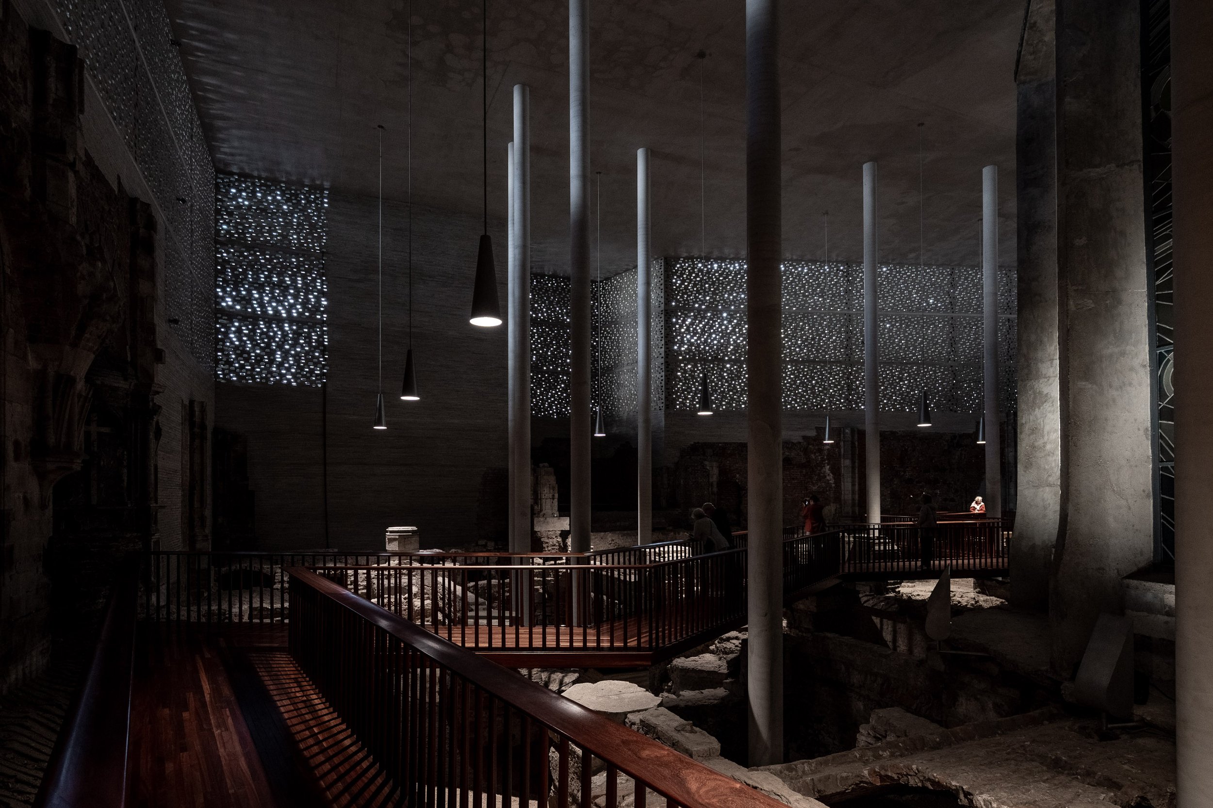 Innenansicht einer archäologischen Stätte, vermutlich einer antiken römischen Ruine, mit erhöhten Holzbrücken und modernen Lichtinstallationen, die eine bedrückende Atmosphäre schaffen.