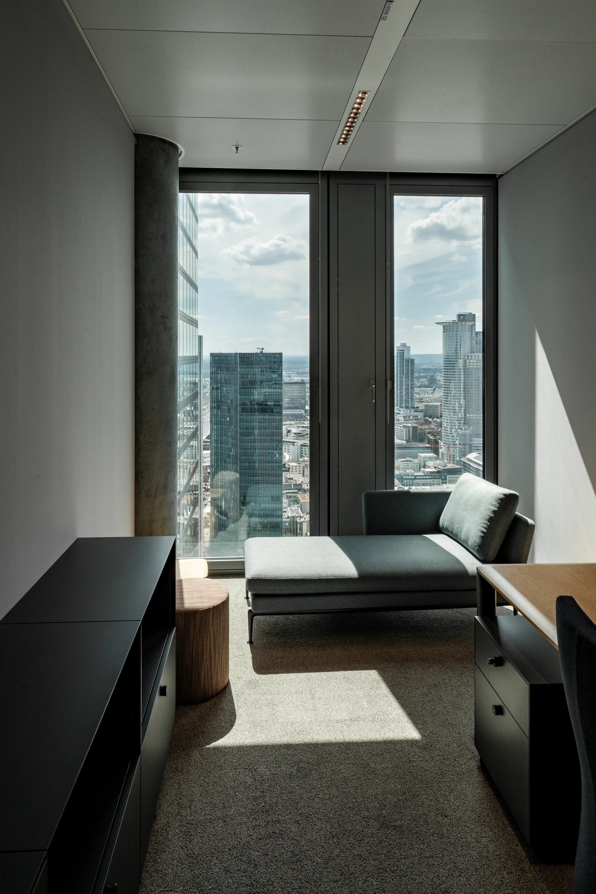 Ein modernes, möbliertes Büro mit Blick auf eine Stadt, ein graues Sofa, eine schwarze TV- oder Konsoleinheit, ein runder Hocker und ein Schreibtisch.