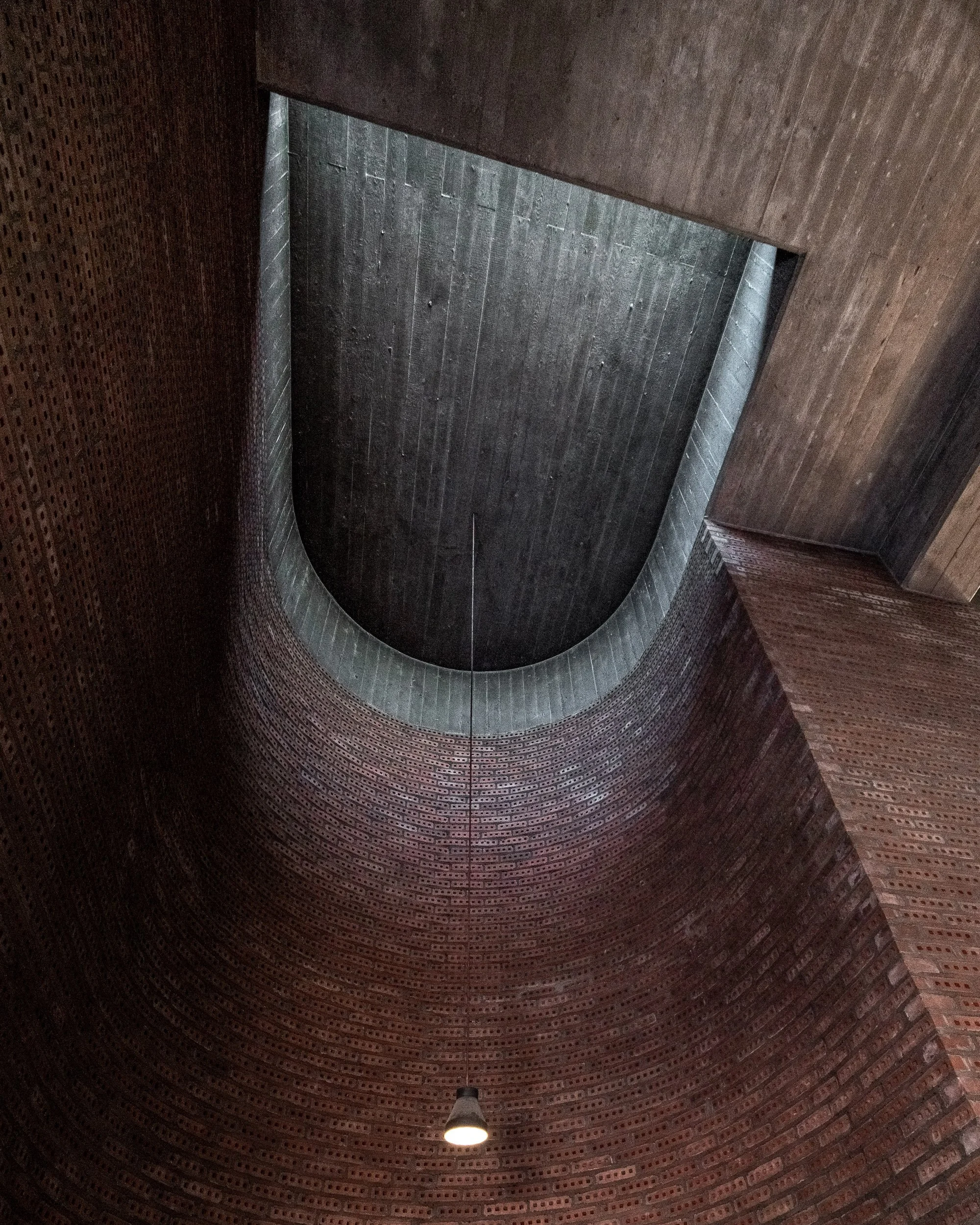 Innenraum eines Gebäudes mit kreisförmiger roter Backsteinmauer, oben ein großes quadratisches Loch im Dach, und eine einzelne Deckenlampe. Blick nach oben.