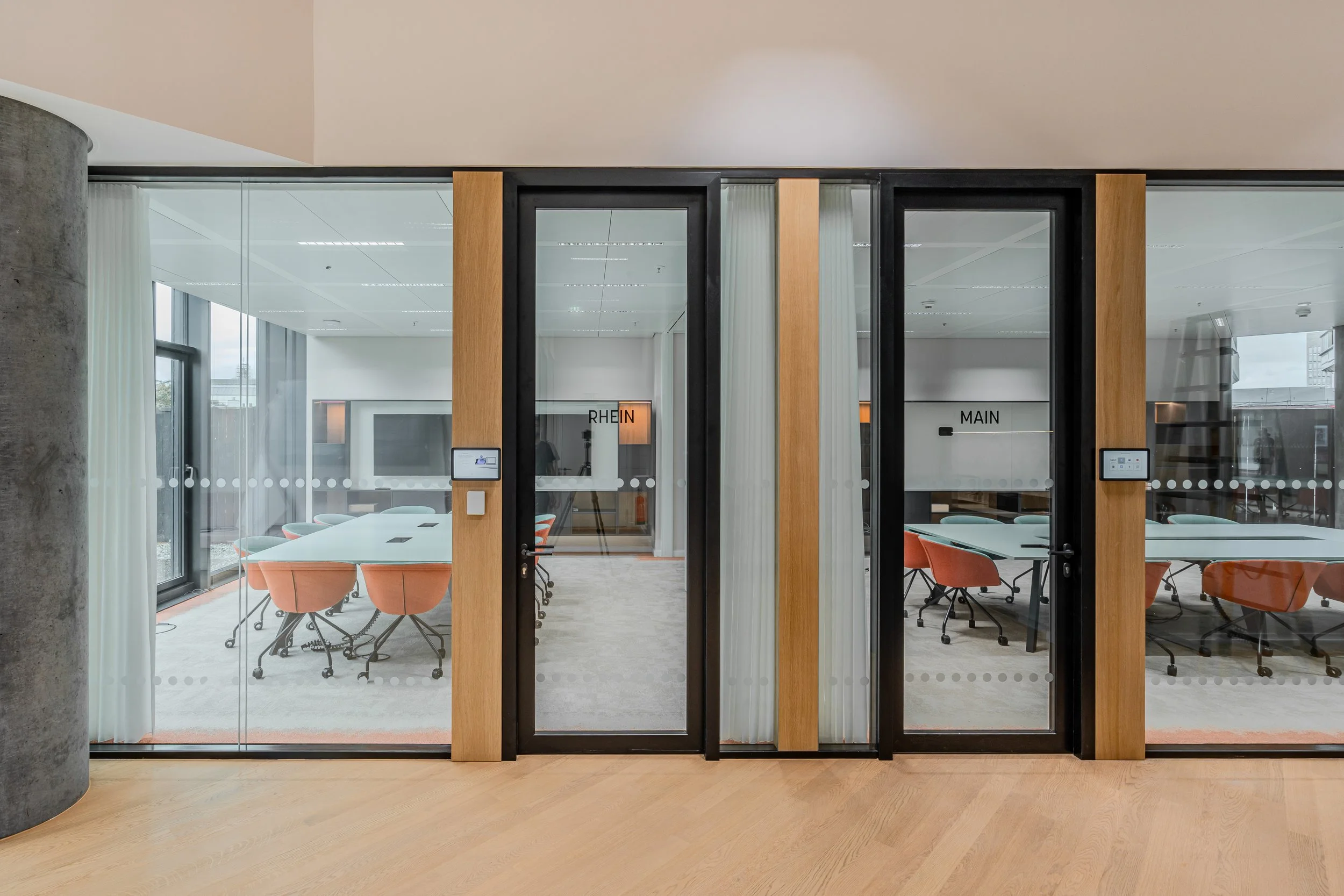 Innenansicht eines modernen Sitzungszimmers mit Glaswänden, schwarzen Rahmen, Holzakzenten, weißen Tischen und orangefarbenen Stühlen.