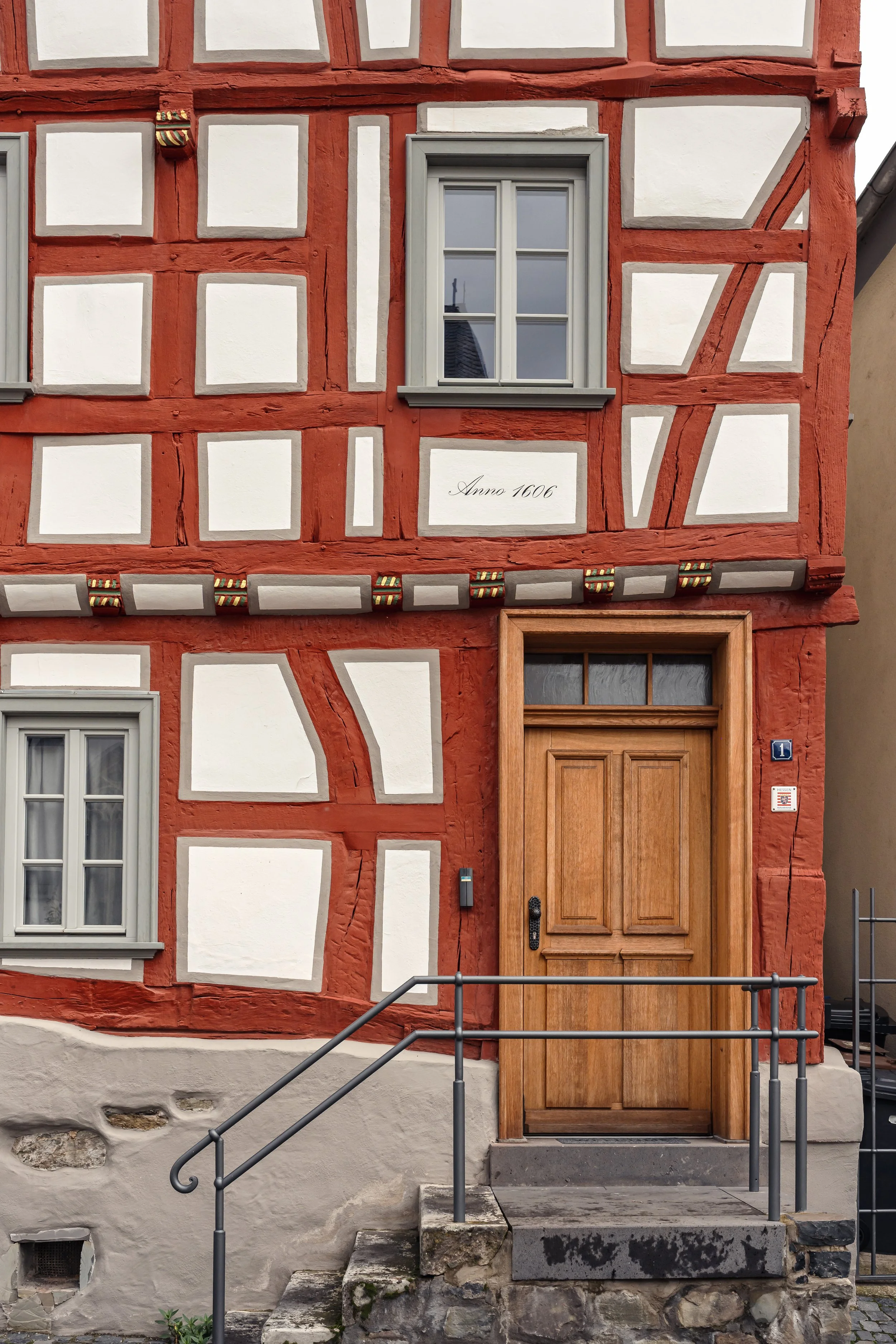 Traditionelles Fachwerkhaus mit rotem Gerüst, weißen Zwischenräumen, grauen Fenstern und einer Holztür. Es trägt die Jahreszahl 'Anno 1606'.