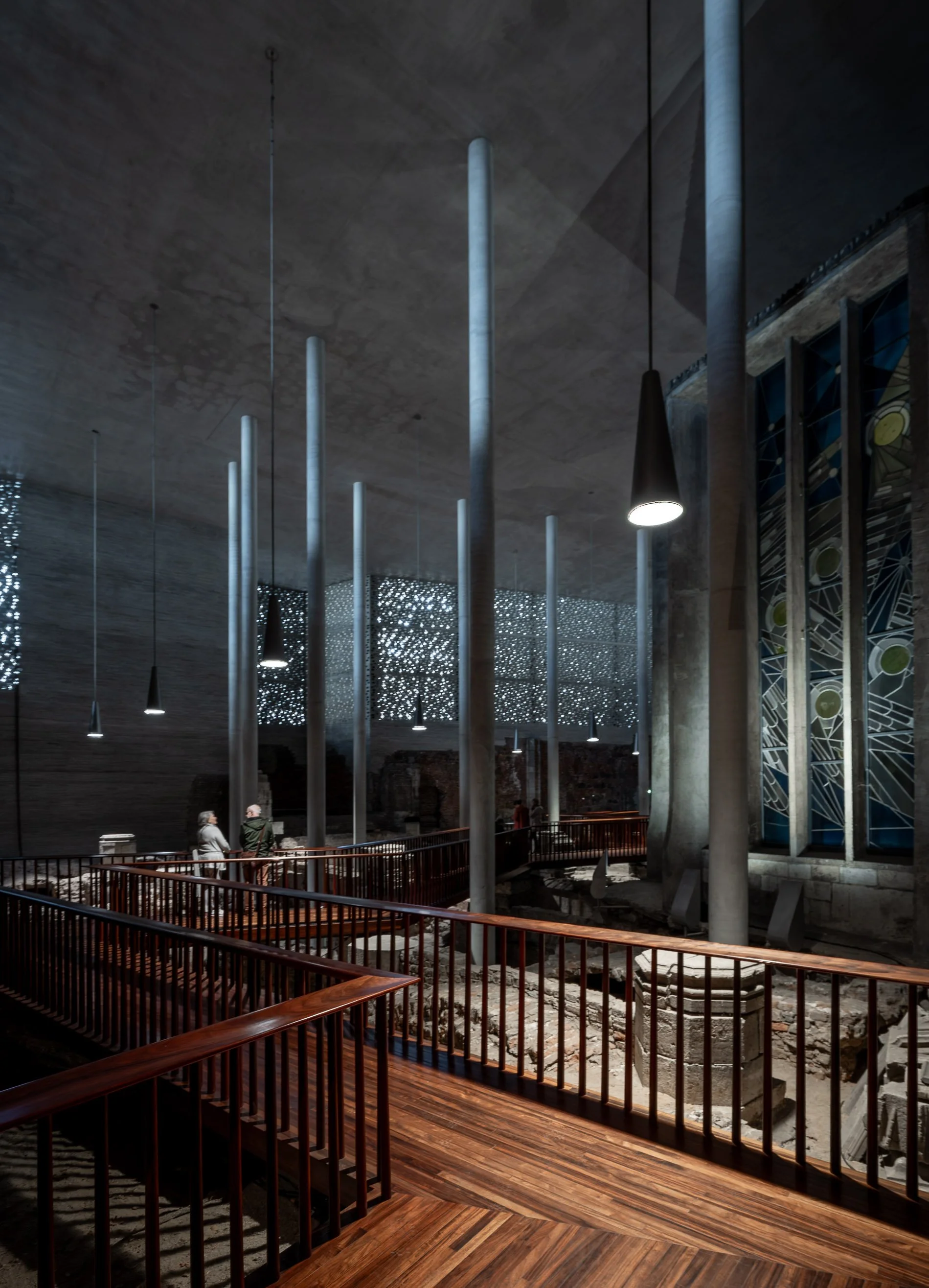 Innenraum eines modernen Gebäudes mit hohen Säulen, Holzboden, dunklen Wänden, modernen Beleuchtungskörpern und einem Fenster mit buntem Glasdesign im Hintergrund.