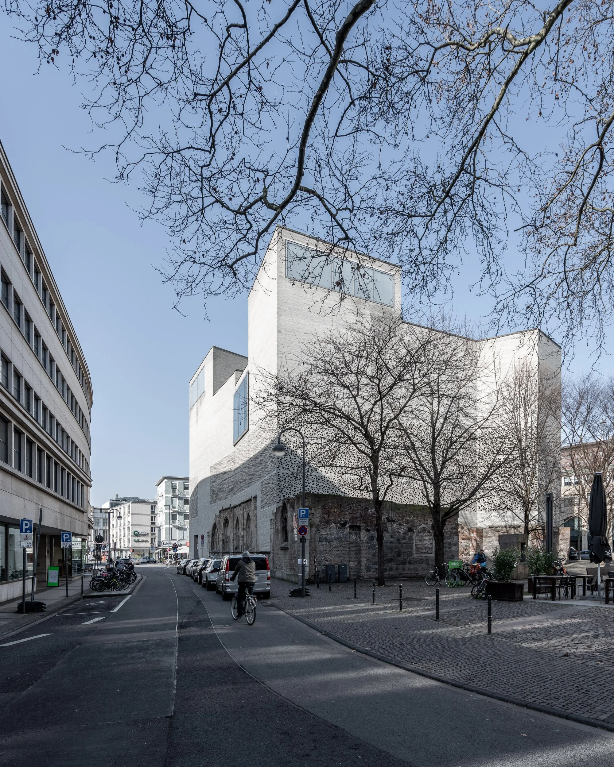 Modernes Gebäude in einer urbanen Straße mit Fahrrädern und Parkplätzen, kahle Bäume im Vordergrund, blauer Himmel
