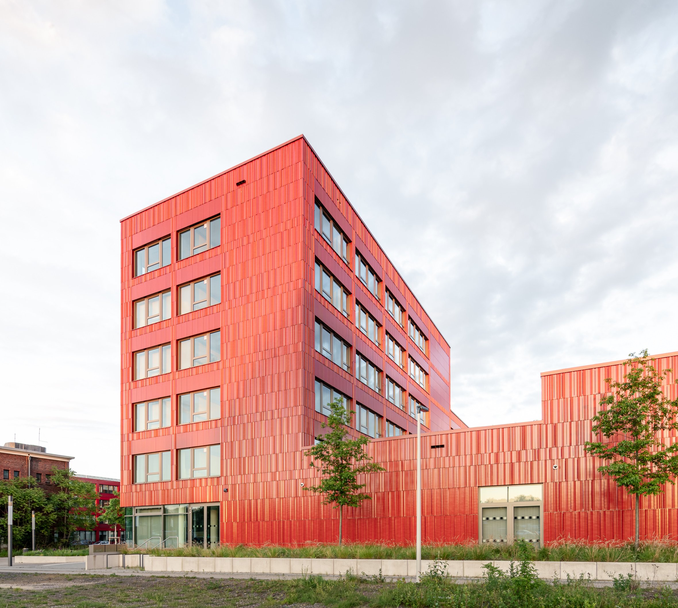 Rotes modernes Gebäude mit vielen Fenstern und vertikaler Fassadengestaltung, umgeben von Grünflächen und Bäumen, unter bewölktem Himmel.