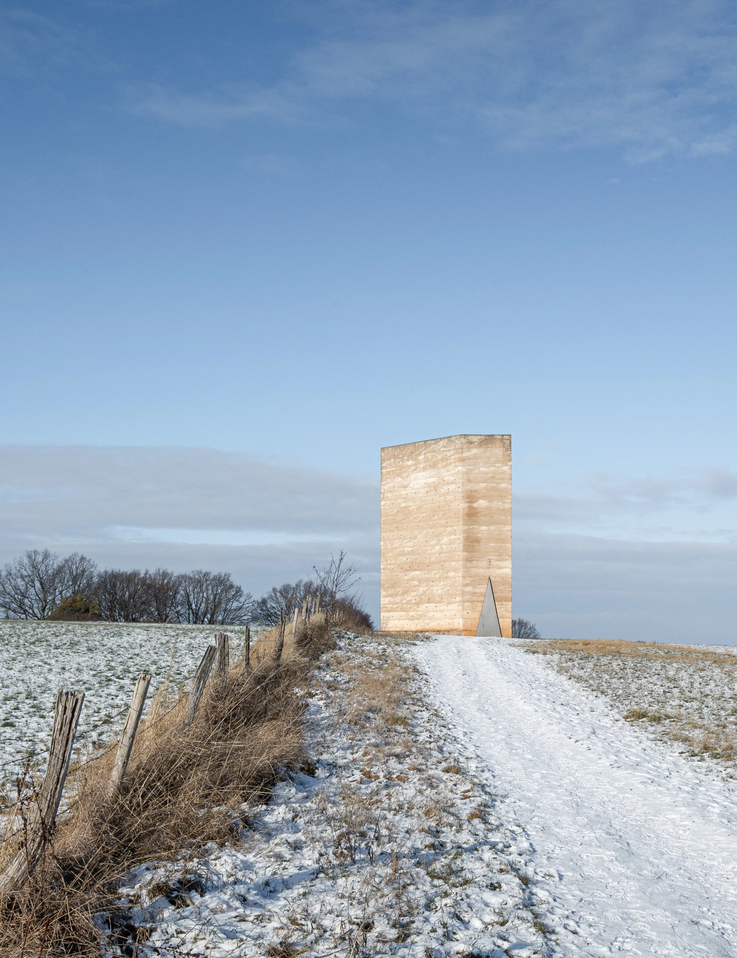 Ein schneebedeckter Weg führt zu einem modernen, rechteckigen Holzgebäude in einer offenen Landschaft mit Bäumen im Hintergrund und einem blauen Himmel.