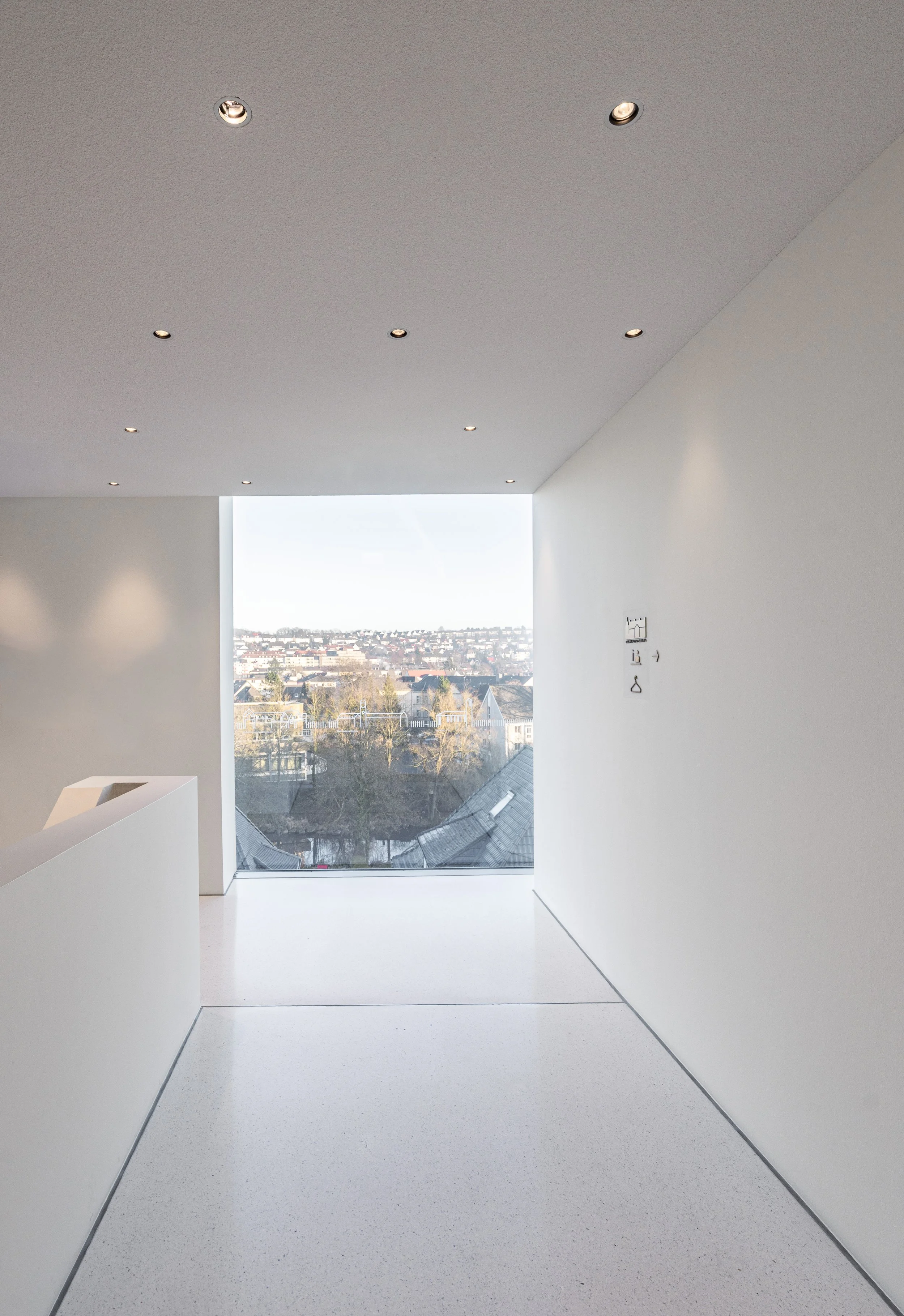 Innenaufnahme eines modernen, hellen Raumes mit großem Fenster, das eine Stadtlandschaft zeigt. Weiße Wände und Decke, kleine Deckenleuchten, minimalistische Gestaltung.