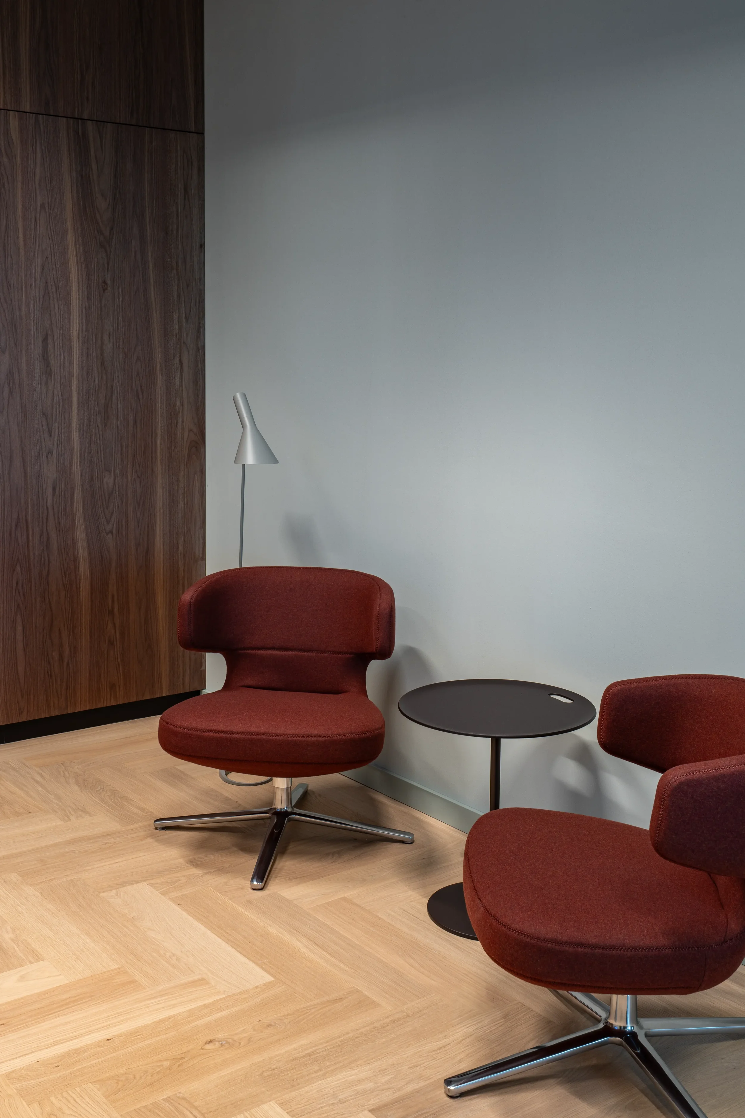 Zwei rote, moderne Sessel mit einem schwarzen Beistelltisch zwischen ihnen, eine Stehlampe im Hintergrund, eine Holz- und eine weiße Wand, Holzboden.