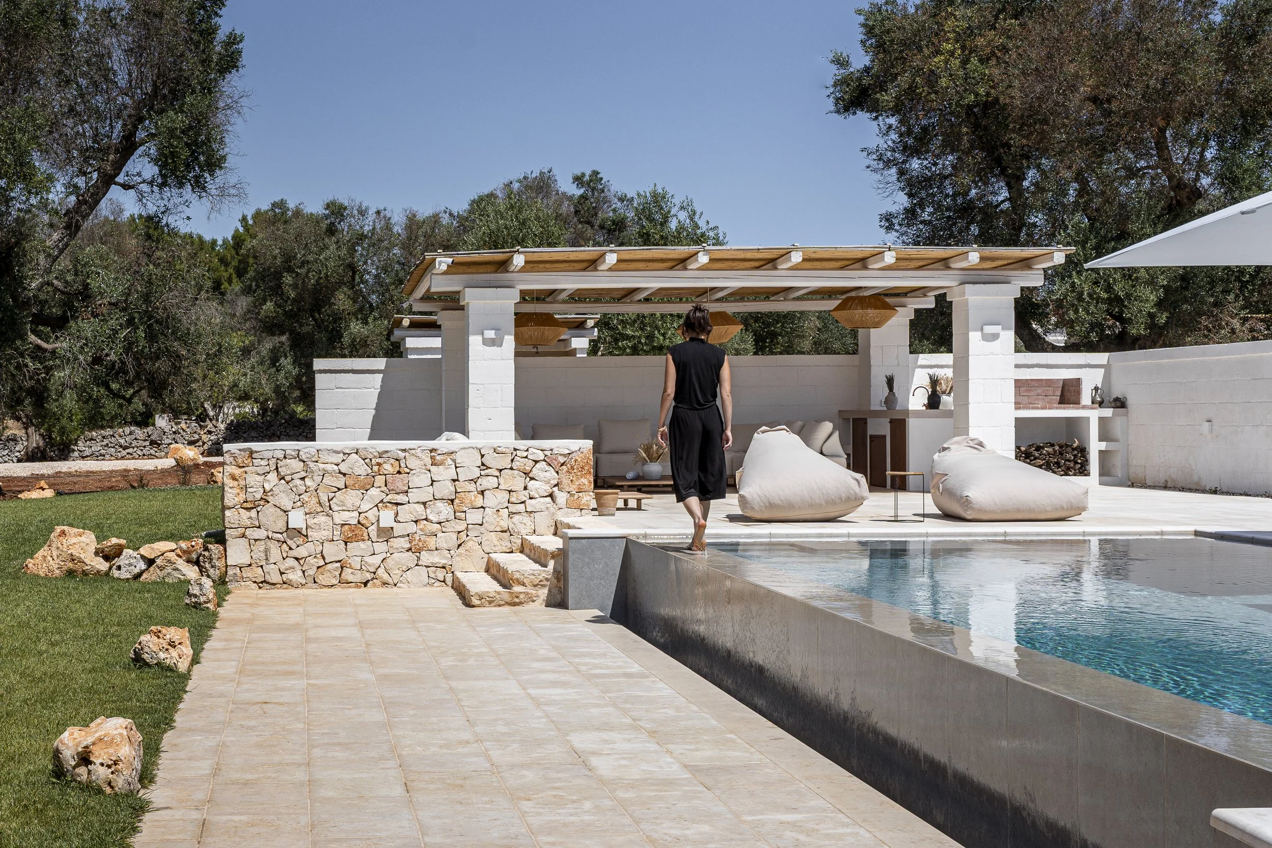 Eine Frau steht an einem Pool im Außenbereich eines modernen Anwesens mit weißen Wänden, Sandsteinboden und einer Sitzecke mit bequemen Sitzsäcken unter einem Holzdach, im Hintergrund Bäume und blauer Himmel.