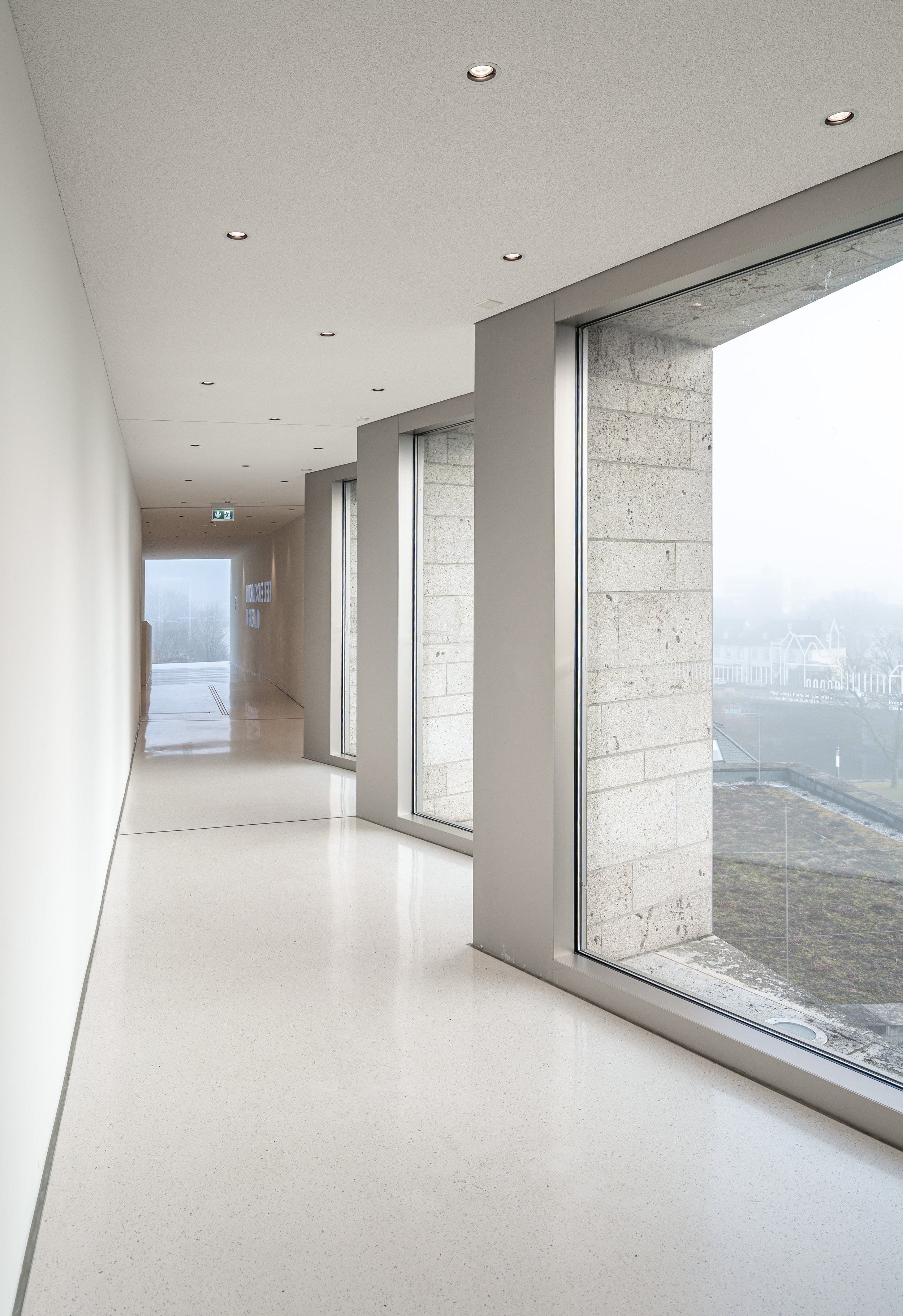 Ein heller, moderner Flur mit großen Fenstern, Blick nach draußen auf eine neblige Landschaft, weiße Wände, glatte weiße Bodenfliesen und eingebaute Deckenleuchten.
