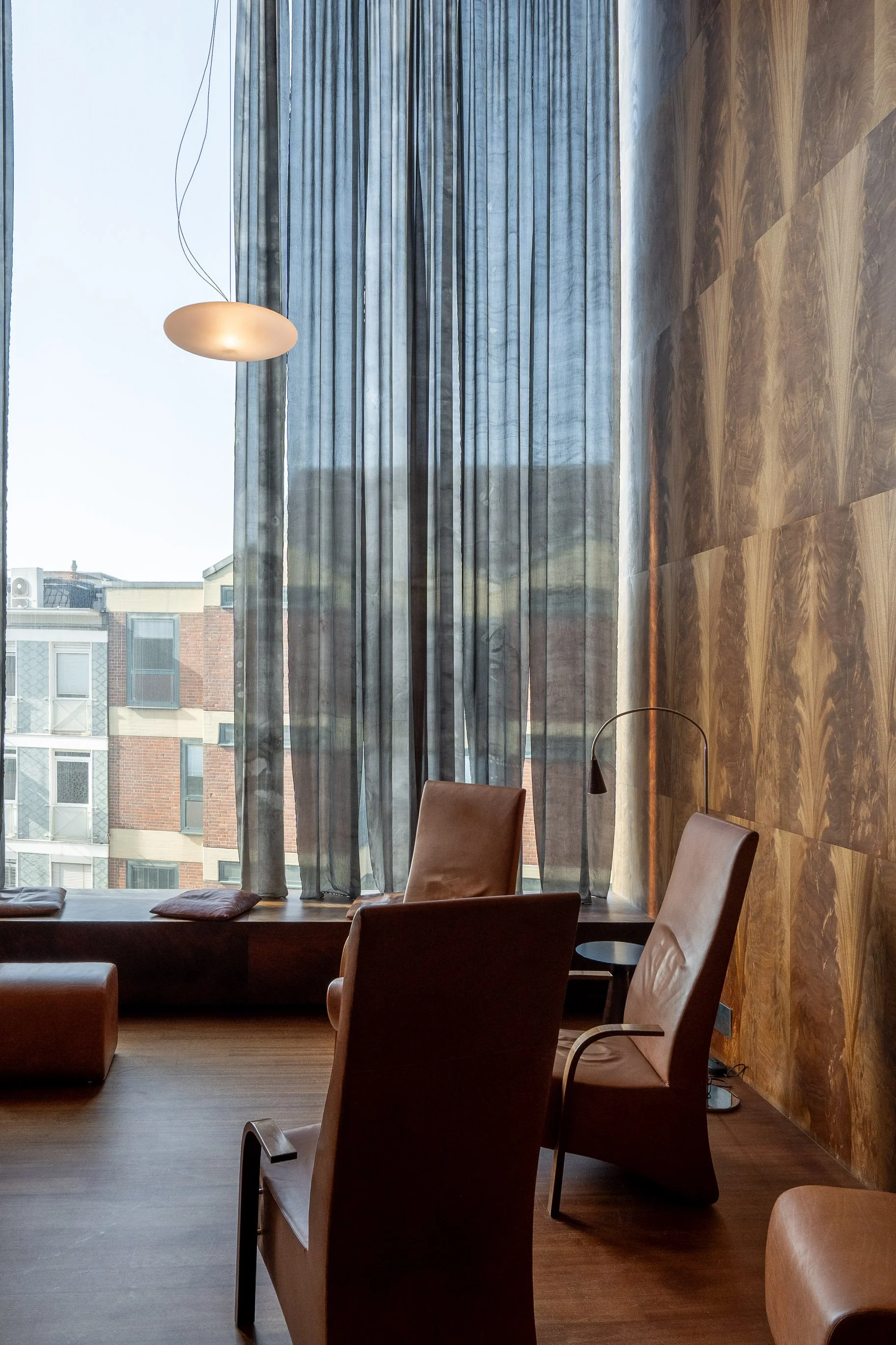 Ein modern eingerichteter Raum mit großen Fenstern, grauen Vorhängen, braunen Sessel, einem kleinen schwarzen Beistelltisch, einer Deckenlampe und einer Wandverkleidung aus Holz.