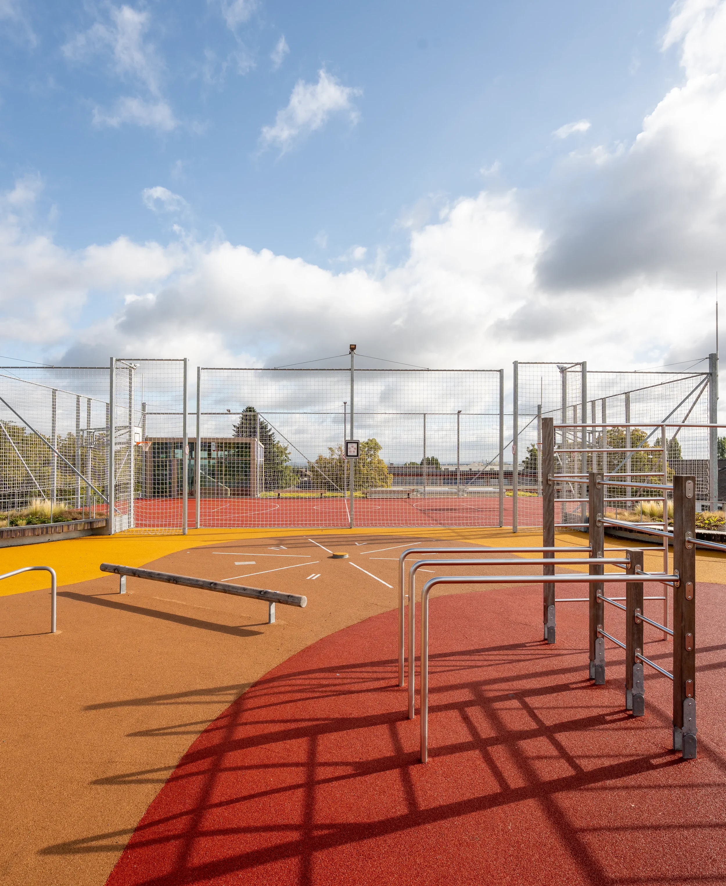 Ein leerer Outdoor-Sportplatz mit Basketballkorb, Turnstangen und Laufbahn, umgeben von einem hohen Netz. Der Himmel ist teilweise bewölkt.