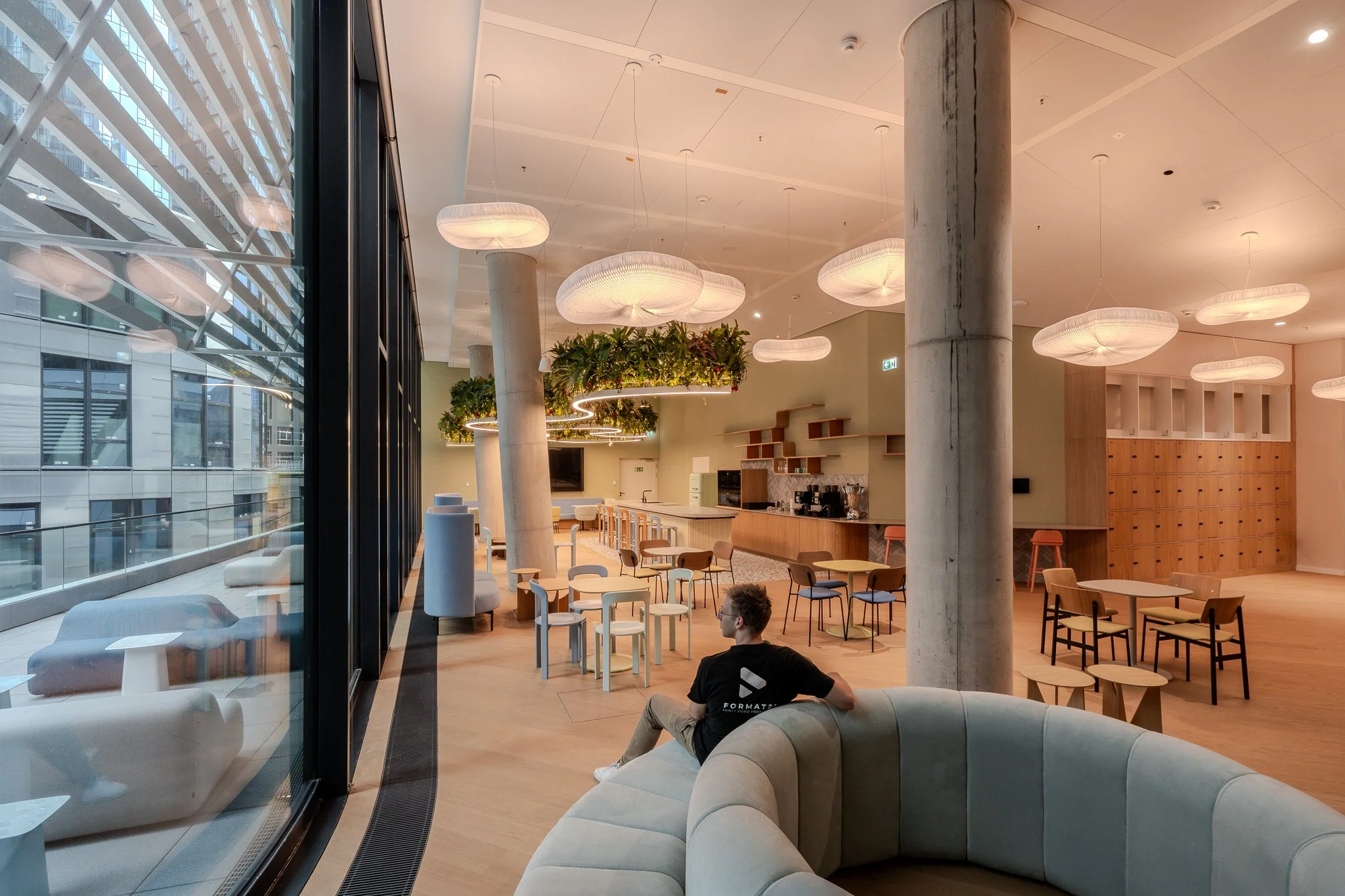 Innenansicht eines modernen, offenen Gemeinschaftsraums mit viel natürlichem Licht, gepolsterten Sitzmöbeln, mehreren kleinen Tischen und Stühlen, großen weißen Deckenleuchten, einem Konferenztisch, einer Küchenzeile sowie einem jungen Mann, der auf 