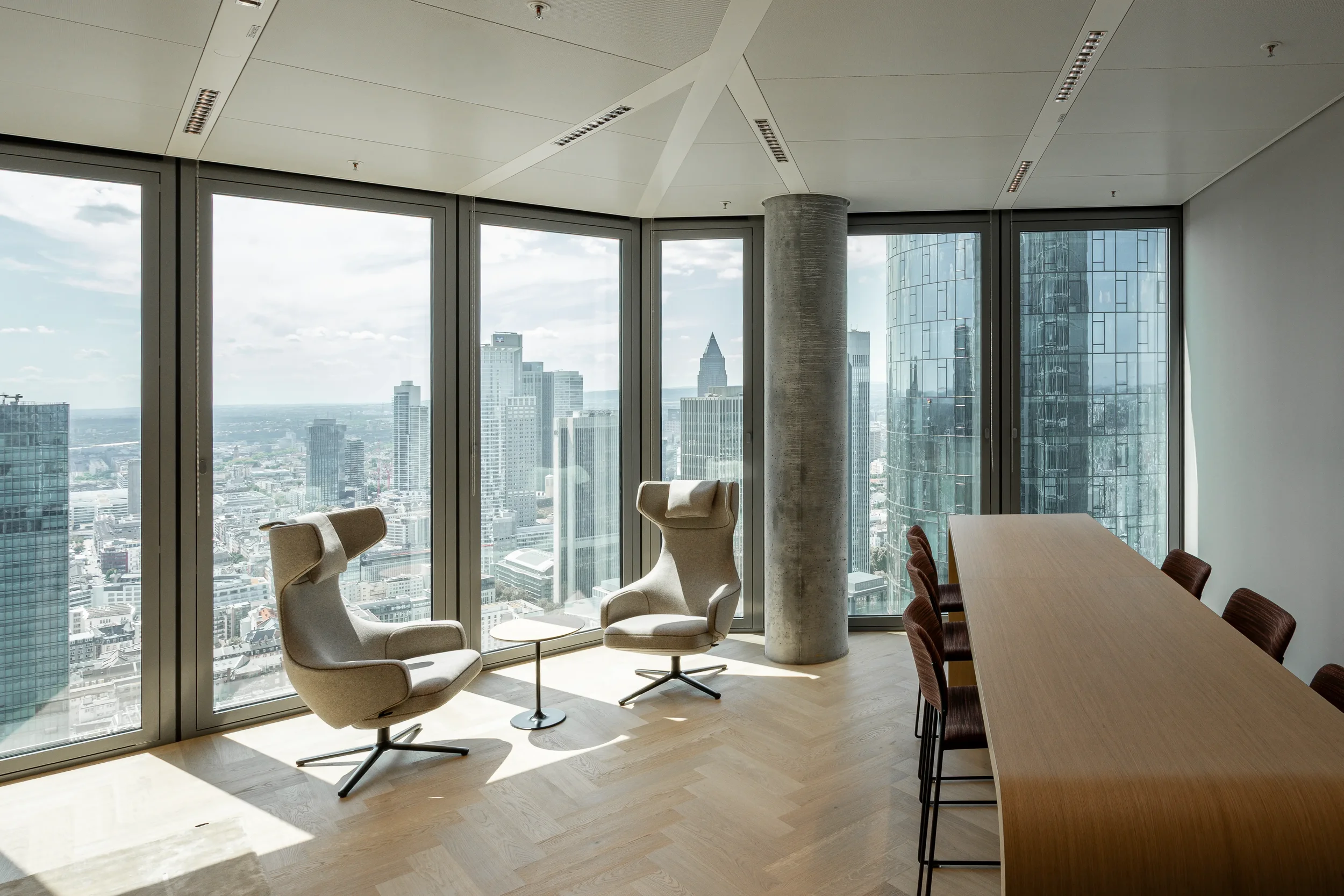 Modernes Büro mit großen Fenstern, City-View, zwei ergonomische Stühle, kleinem Tisch und einem langen Holztisch mit Stühlen.