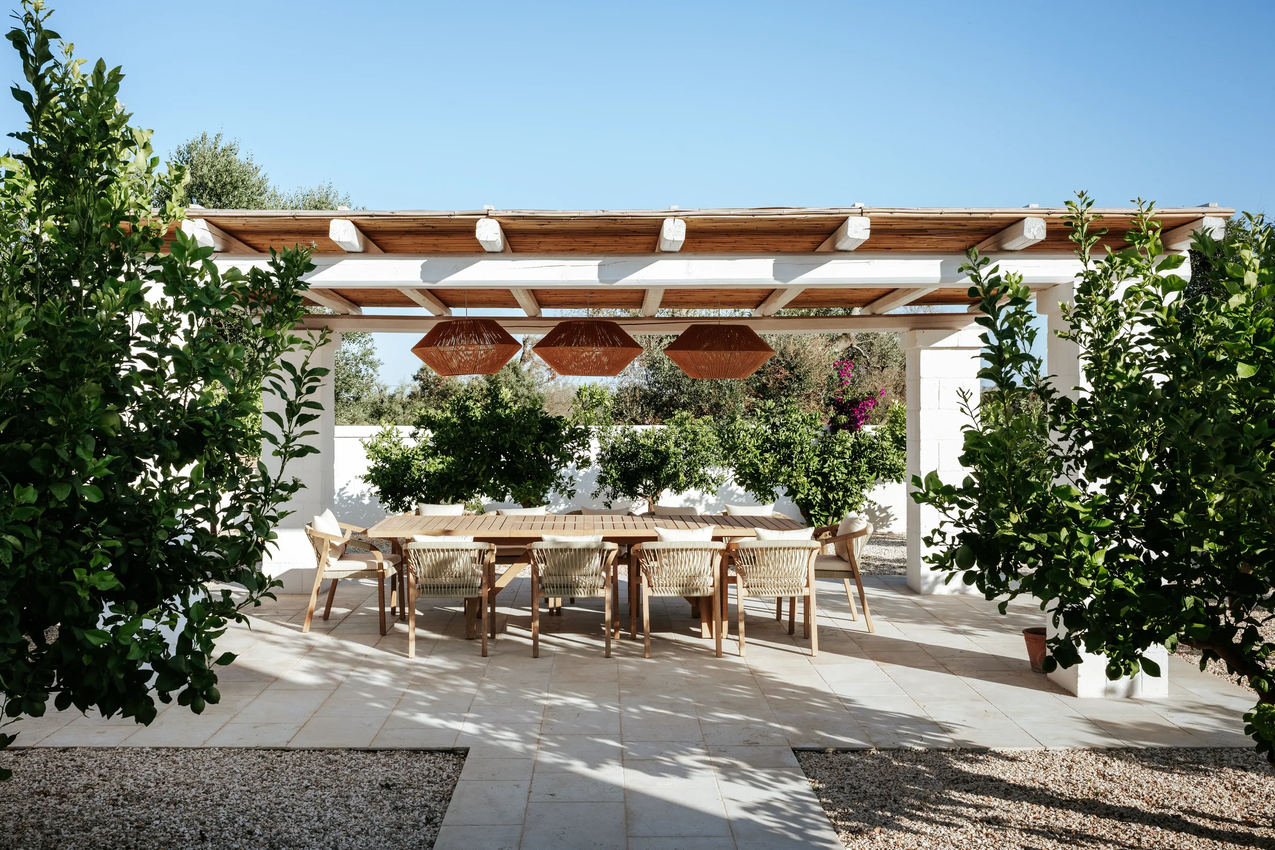 Eine überdachte Terrasse mit einem großen Holztisch und mehreren Stühlen, umgeben von grünen Sträuchern bei sonnigem Wetter.