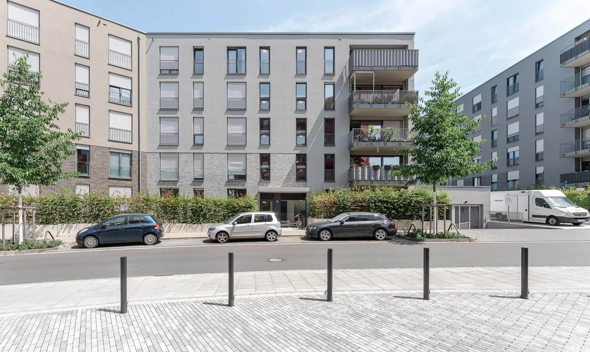 Moderne mehrstöckige Wohngebäude mit Balkonen, parkende Autos und Bäume vor einem Stadtstraßen. Klare, sonnige Tageszeit, Himmel mit wenigen Wolken.