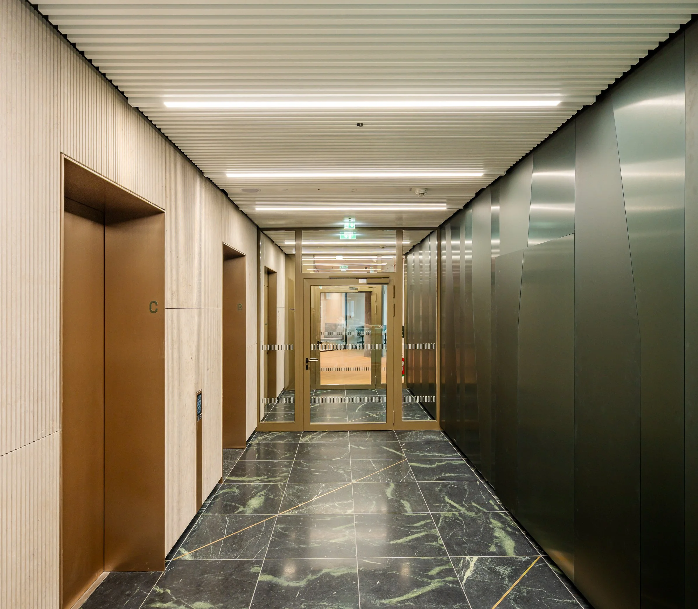 Innenansicht eines modernen Aufzugs in einem Gebäude mit eleganten schwarzen Marmorfliesen am Boden, einer hellen Holzpaneelwand auf der linken Seite und einer Metallwand auf der rechten Seite, mit automatischen Türen und einem Fluchtlicht im Hinterg
