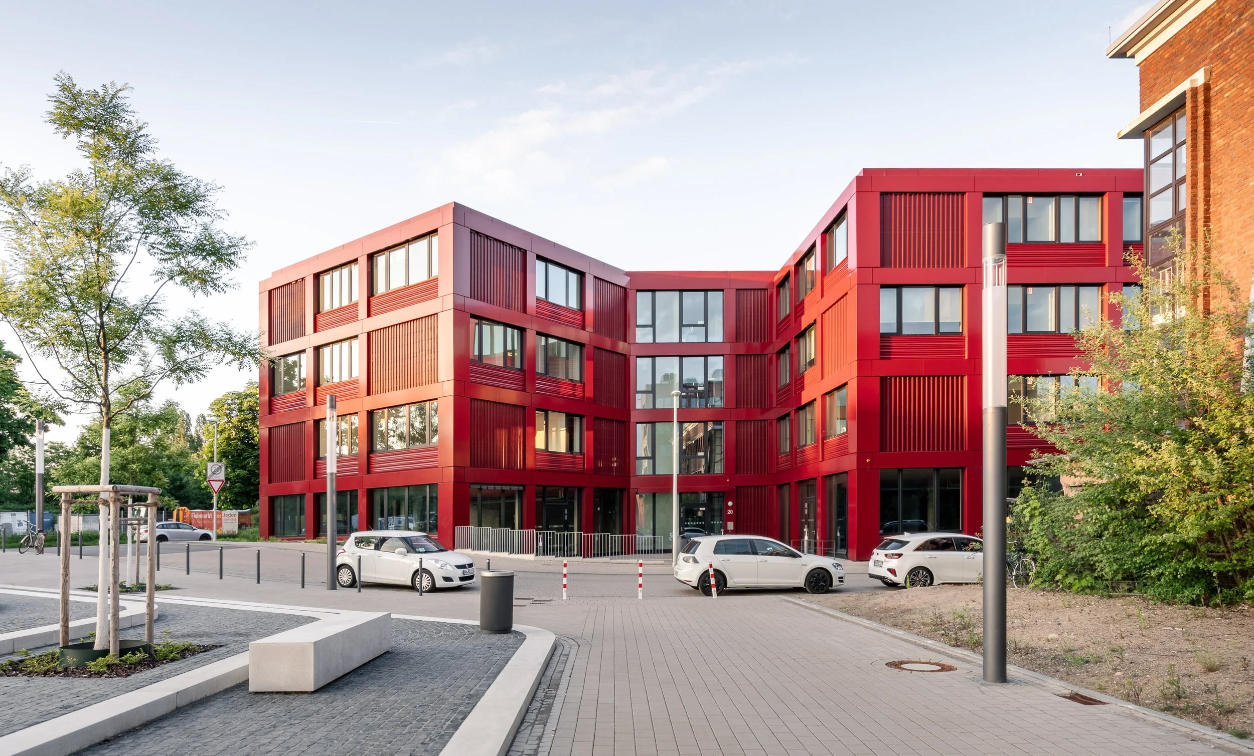 Modernes rotes Gebäude mit großen Fenstern, umgeben von Bäumen und parkenden Autos