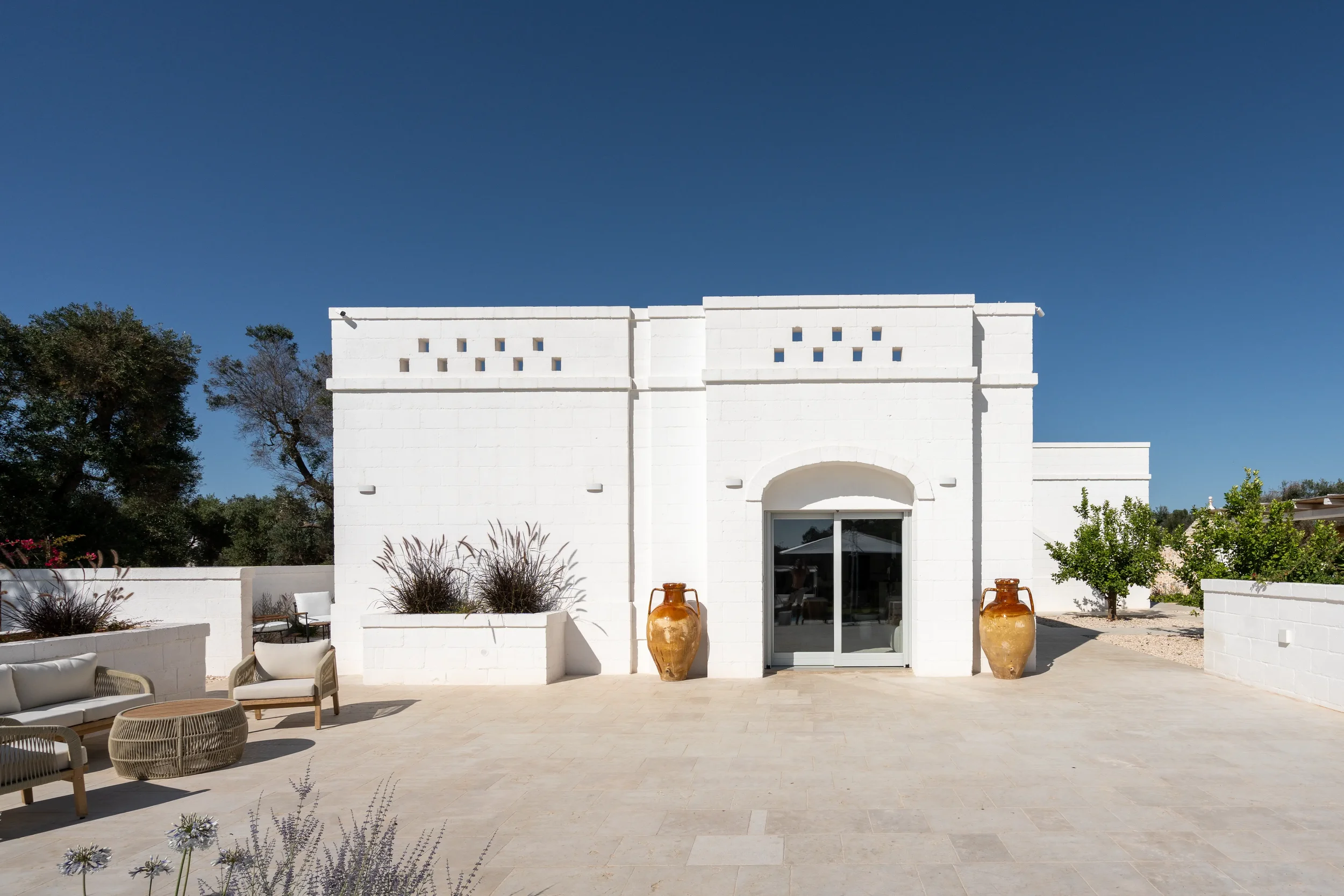 Weiße Pavillon-Architektur vor klarem blauen Himmel, mit Terrasseneinrichtung, großen antiken Töpfen und grünem Gebüsch, umgeben von Natur.