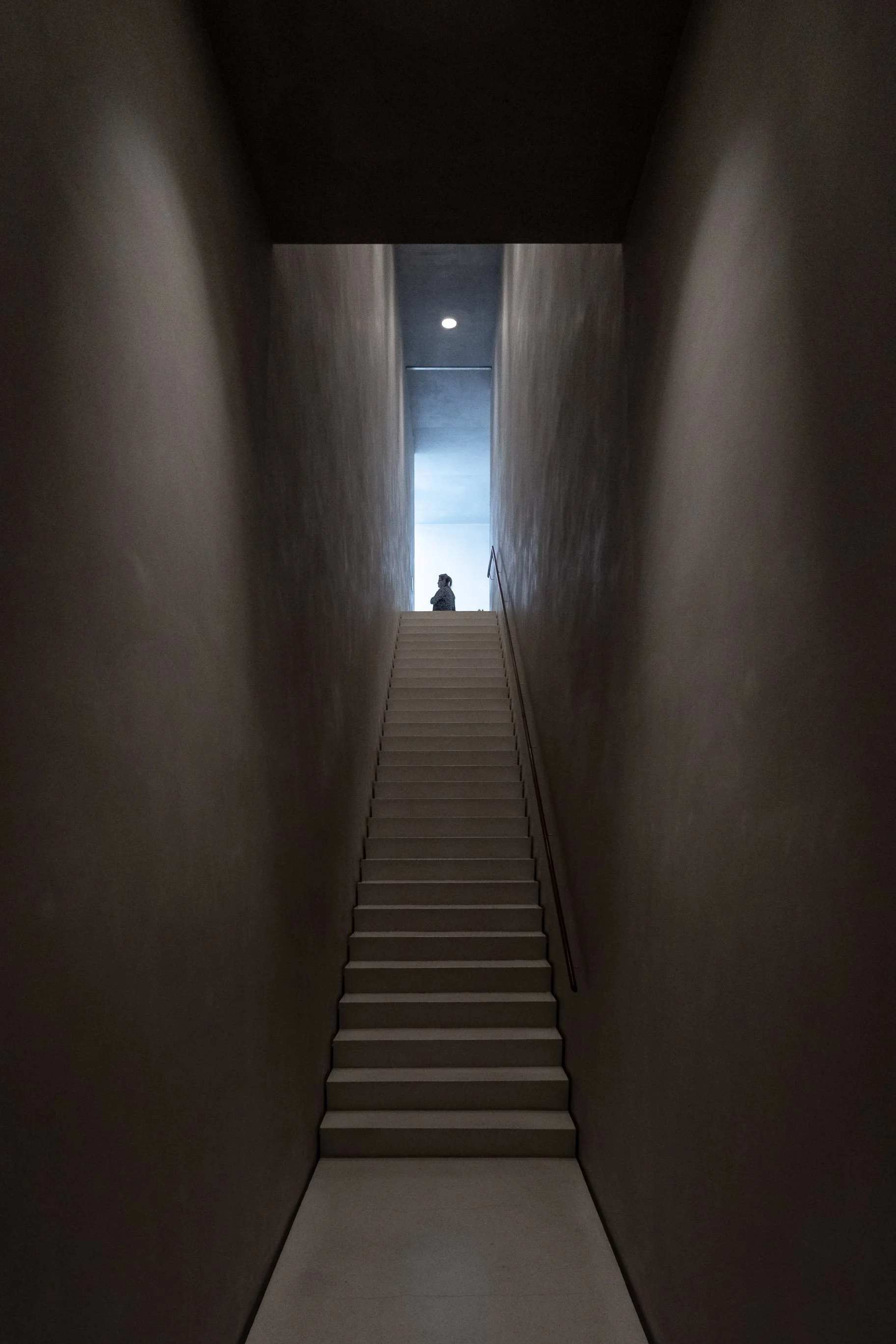 Ein schmaler, dunkler Innenraum mit roten Wänden, der zu einer Treppe führt, die nach oben zu einer hellen Öffnung führt, an der eine Person steht.