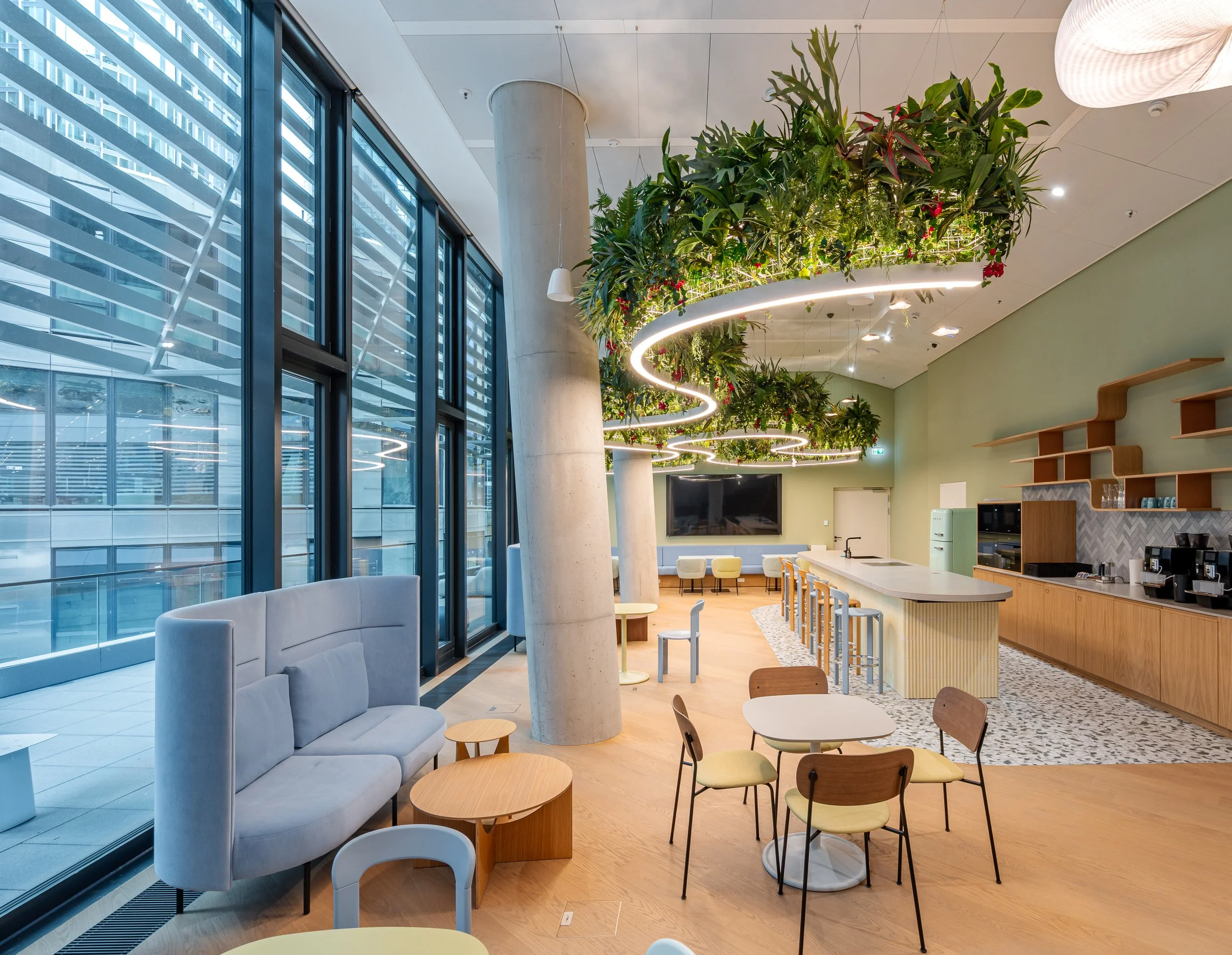 Moderne Cafeteria im FOUR Frankfurt mit Barista-Bereich, großen Fenstern, Pflanzenhängetröge mit Lichteranzug, gemütlichen Sitzgelegenheiten, einem großen Bildschirm, Holzwänden und einem Kaffeeautomaten.