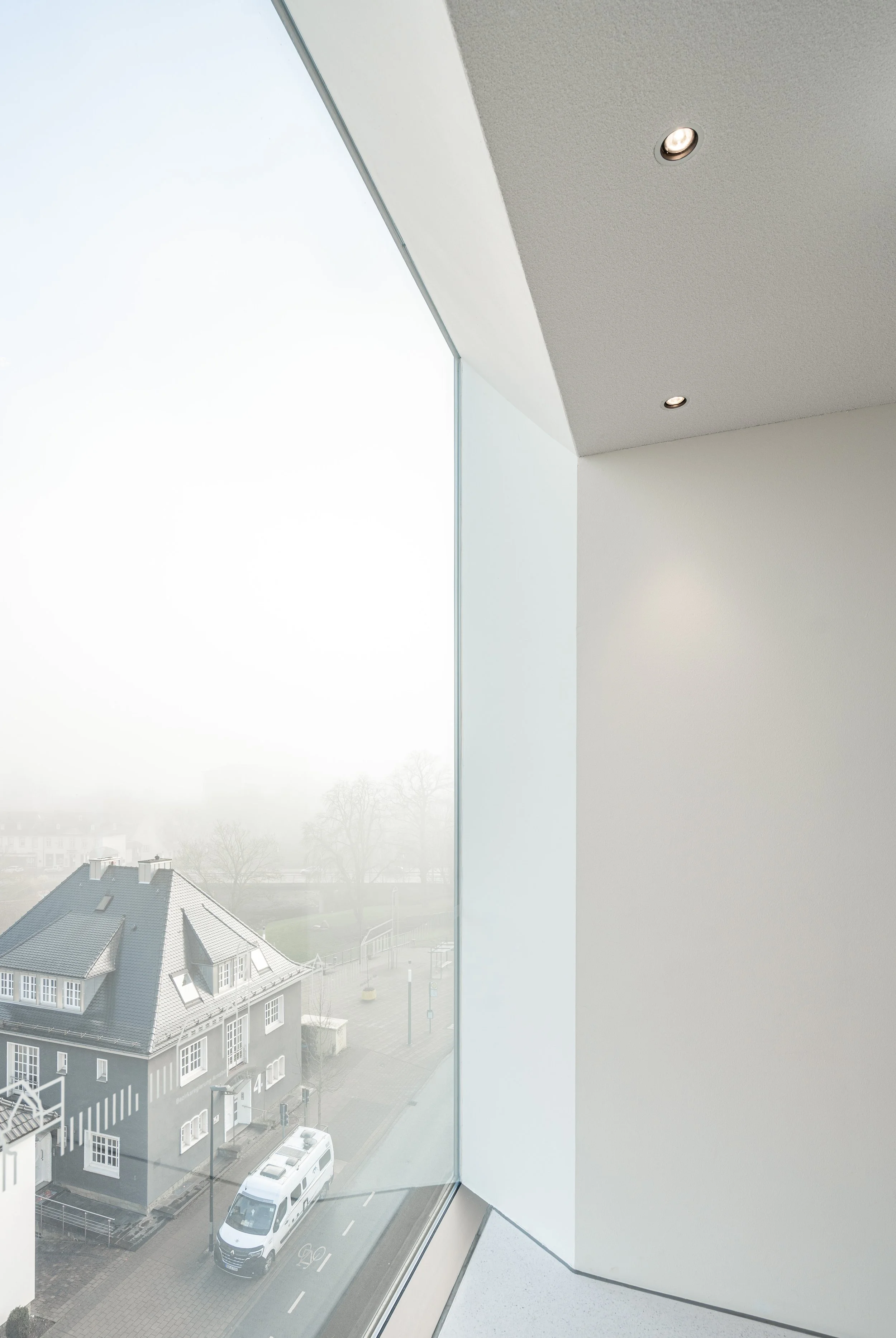 Innenraum mit großen bodentiefen Fenstern, durch die man eine Straße mit einem weißen Van und einem zweistöckigen Haus mit grauem Dach sieht, sowie nebligem Himmel im Hintergrund.