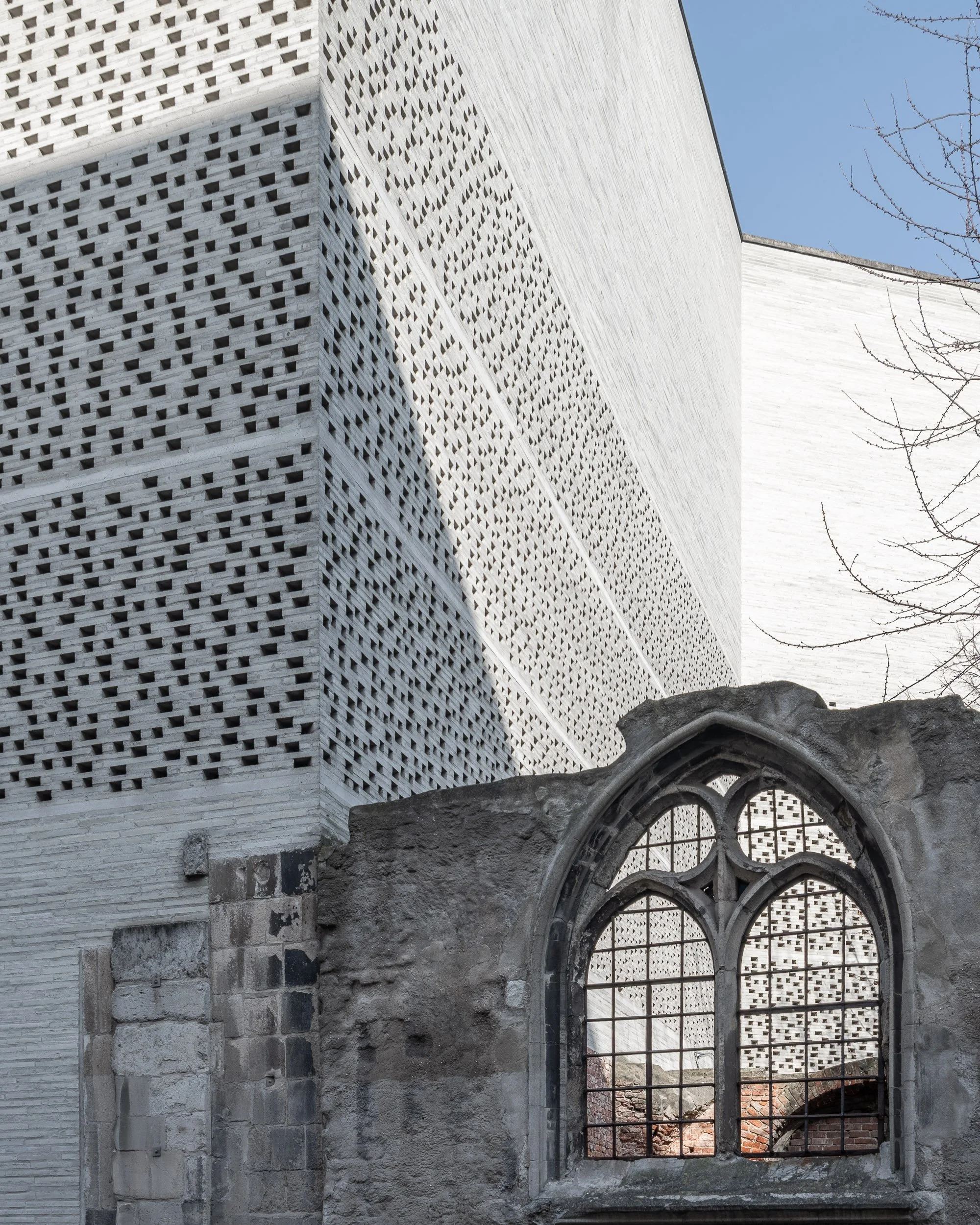 Ein moderner Gebäudeteil mit weißen Wänden und einer dezenten Lochmusterung, sowie ein alteingesessene gotische Fensteröffnung aus Stein, umgeben von einer Mauer, mit Baumästen im rechten Bildbereich. Klare blauer Himmel im Hintergrund.