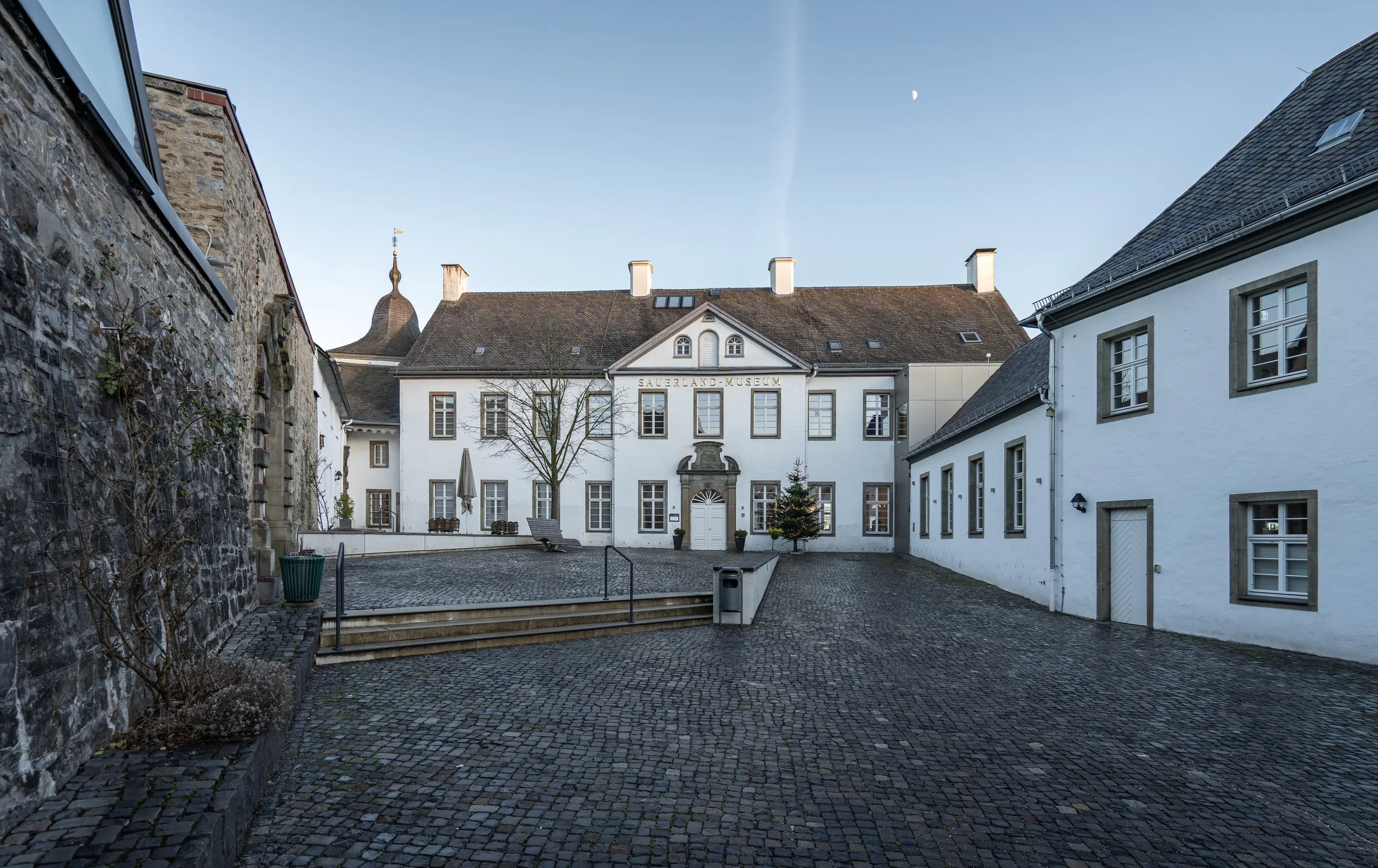 Das Bild zeigt das Sauerland-Museum in einem historischen Gebäude mit weißer Fassade, mehreren Fenstern und einem Eingang. Die umliegende Fläche ist gepflastert. Es gibt einige Bäume, eine Weihnachtsbaumdekoration und einen kleinen Hof.
