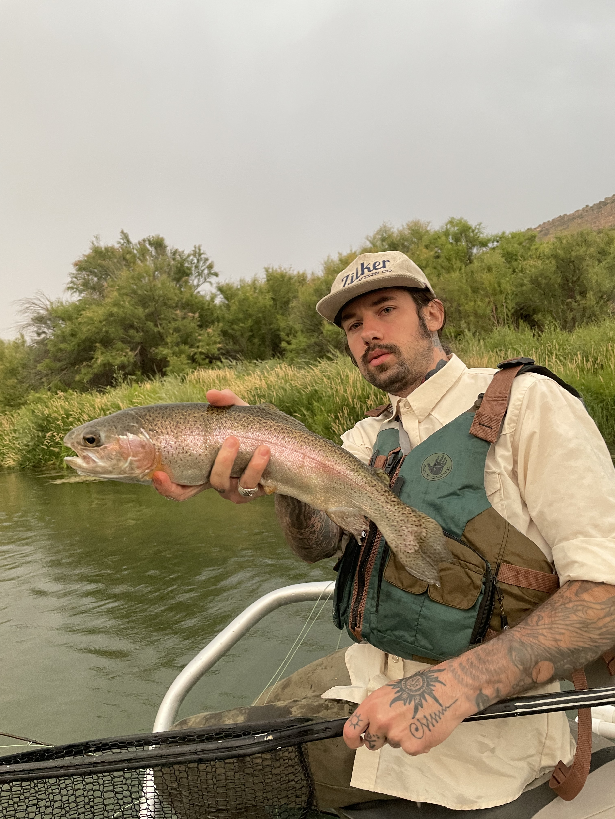 A man holding a large rainbow trout fish on a boat in a river, with grassy and tree-covered hills in the background.