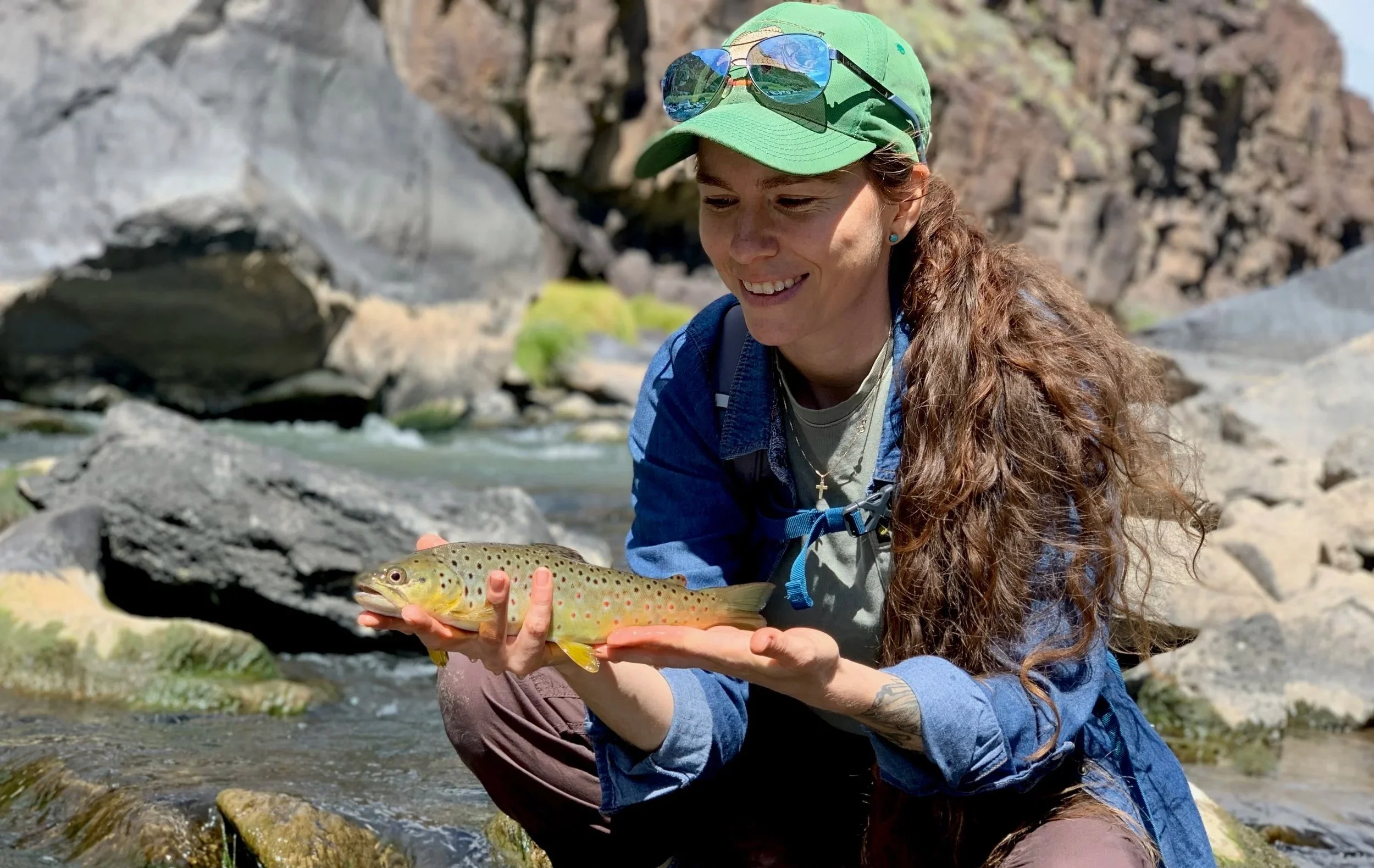 A woman with long curly hair, wearing a green cap and sunglasses, holding a colorful trout fish in a clear mountain stream surrounded by rocks.