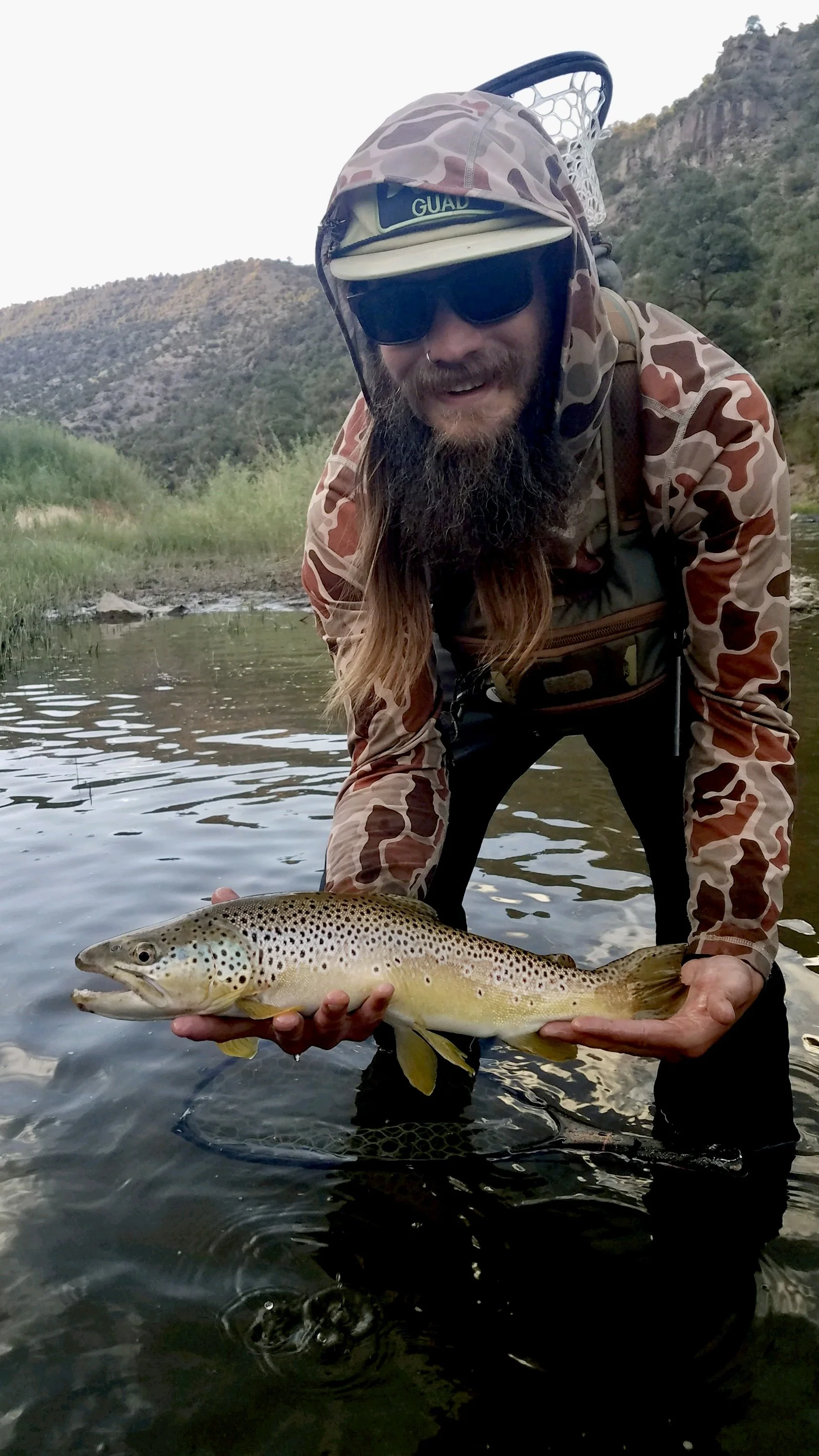 A man with a beard wearing camouflage clothing, sunglasses, and a cap is kneeling in a shallow river holding a large fish, possibly a trout, in both hands. The background shows a mountainous landscape with green trees.