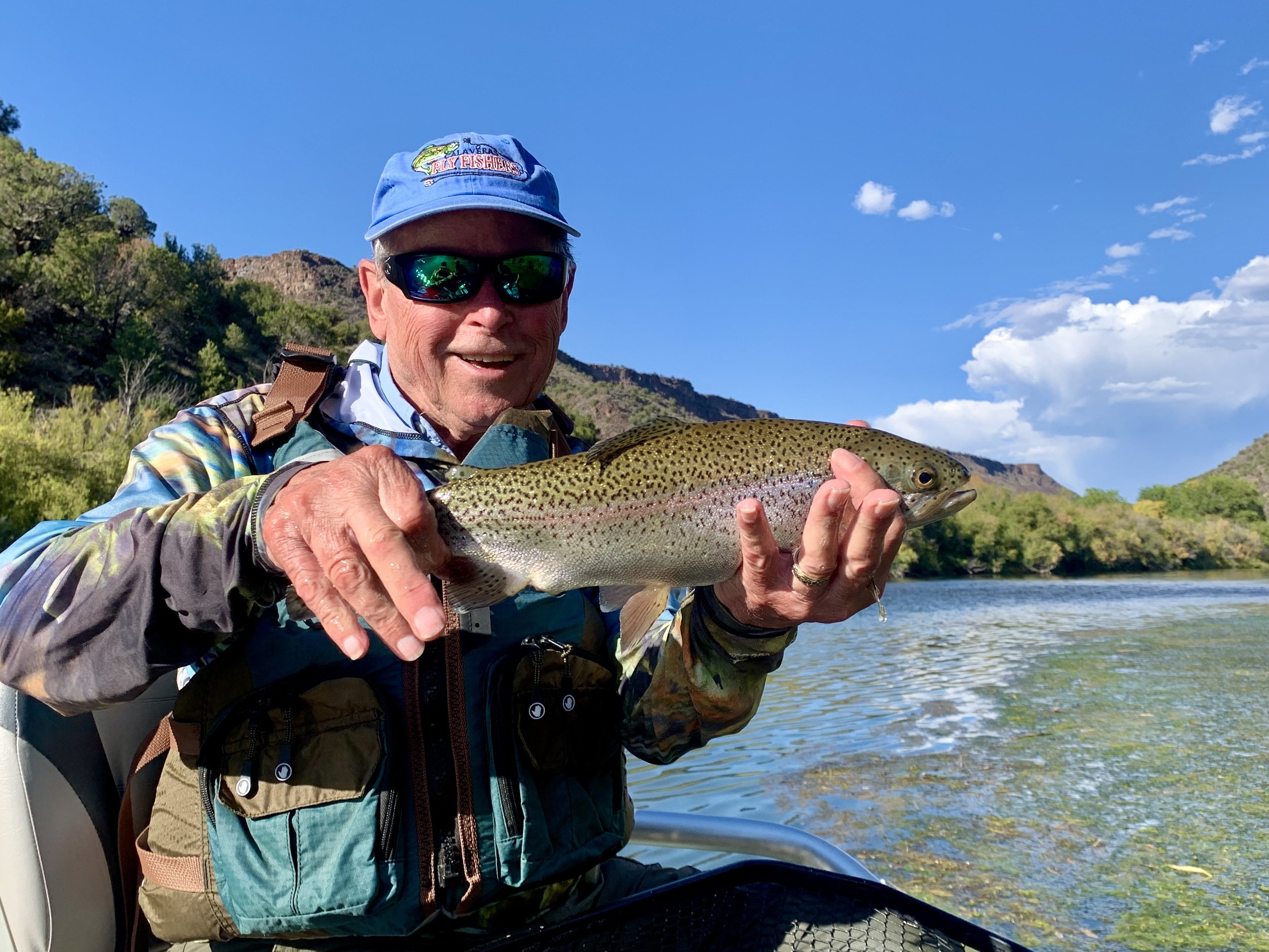 A man wearing sunglasses, a blue cap, and outdoor gear is smiling while holding a fish he caught. The background features water, trees, and mountains under a blue sky with a few clouds.