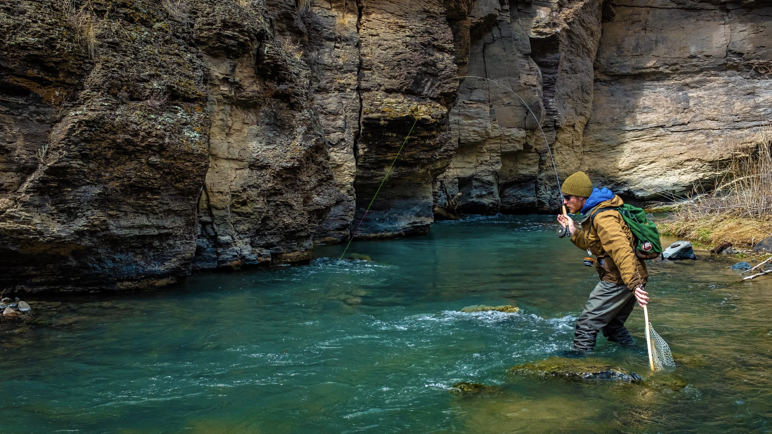 Man in outdoor gear fishing in a river surrounded by rocky cliffs.