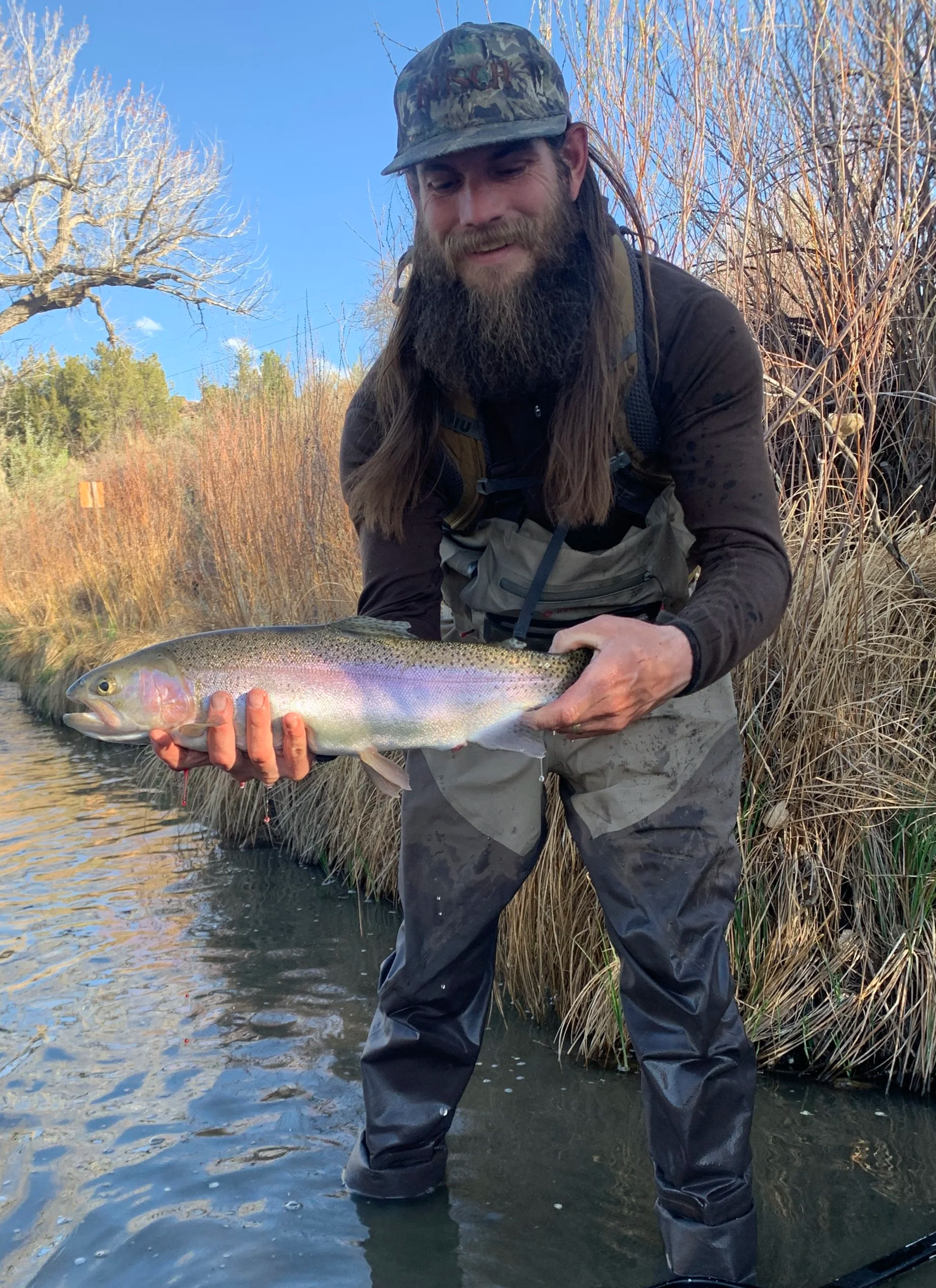 Man with long hair, beard, and camouflage hat holding a large rainbow trout in a creek with dry grass and trees in the background.