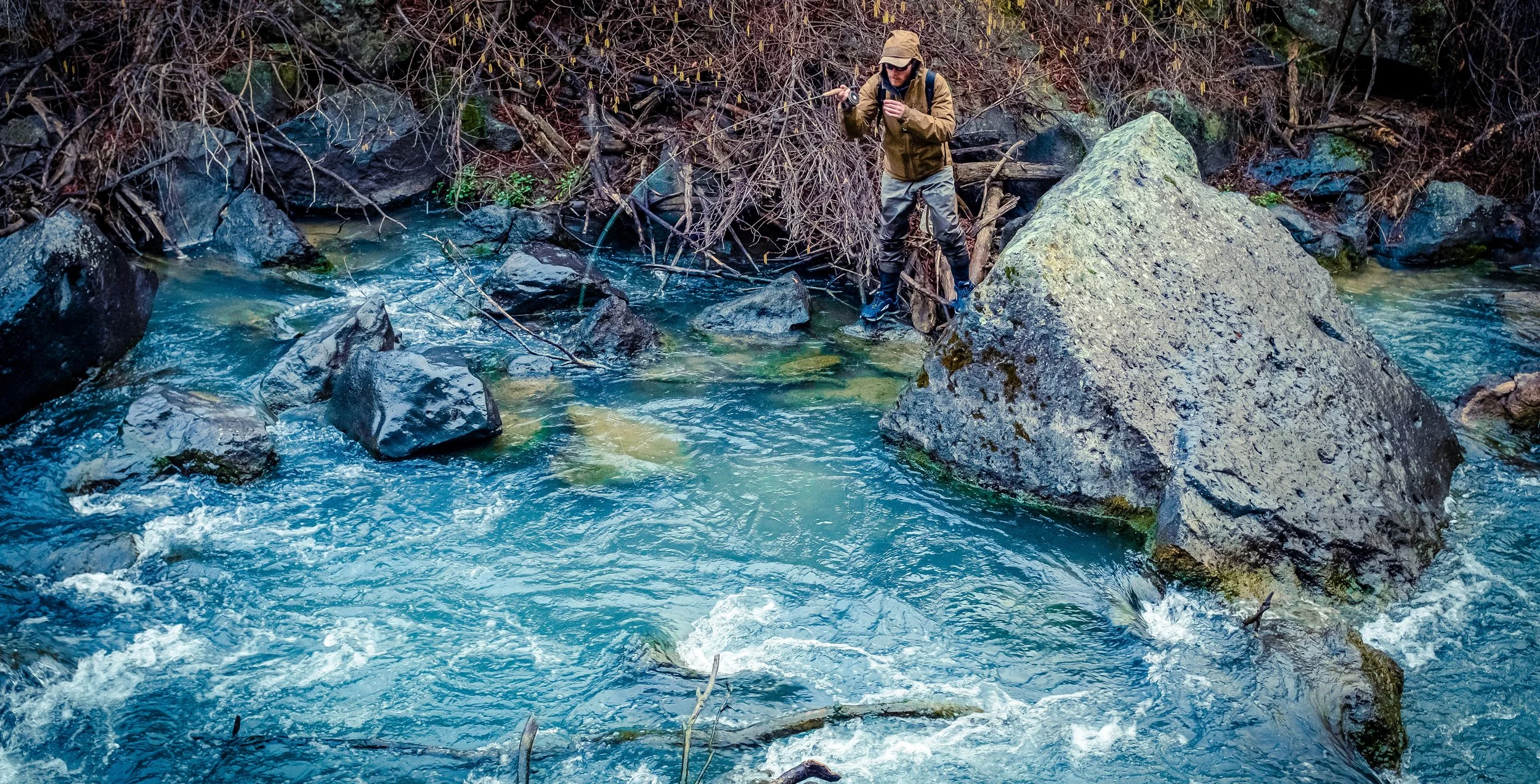 A person in outdoor gear fishing in a rocky river surrounded by bare trees and overhanging branches.