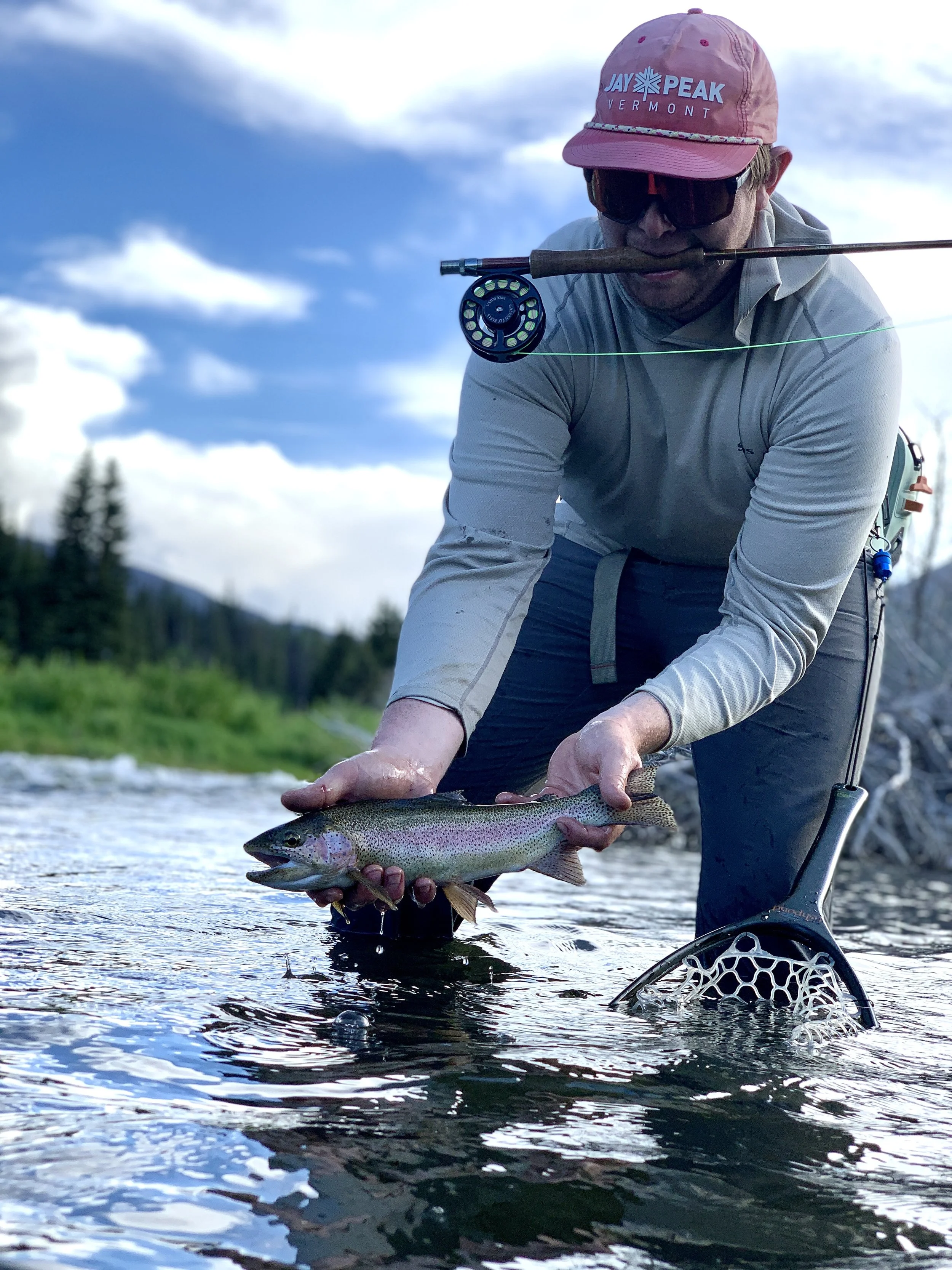 Man in outdoor clothing holding a rainbow trout in a river, wearing a pink hat and sunglasses, with fishing gear nearby.