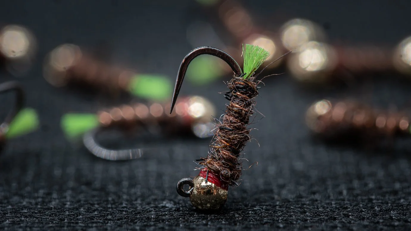 Close-up of a fishing fly with a metal weight at the bottom, wrapped with dark brown thread, and a small green feather at the top, placed on a black textured surface with blurred fishing flies in the background.
