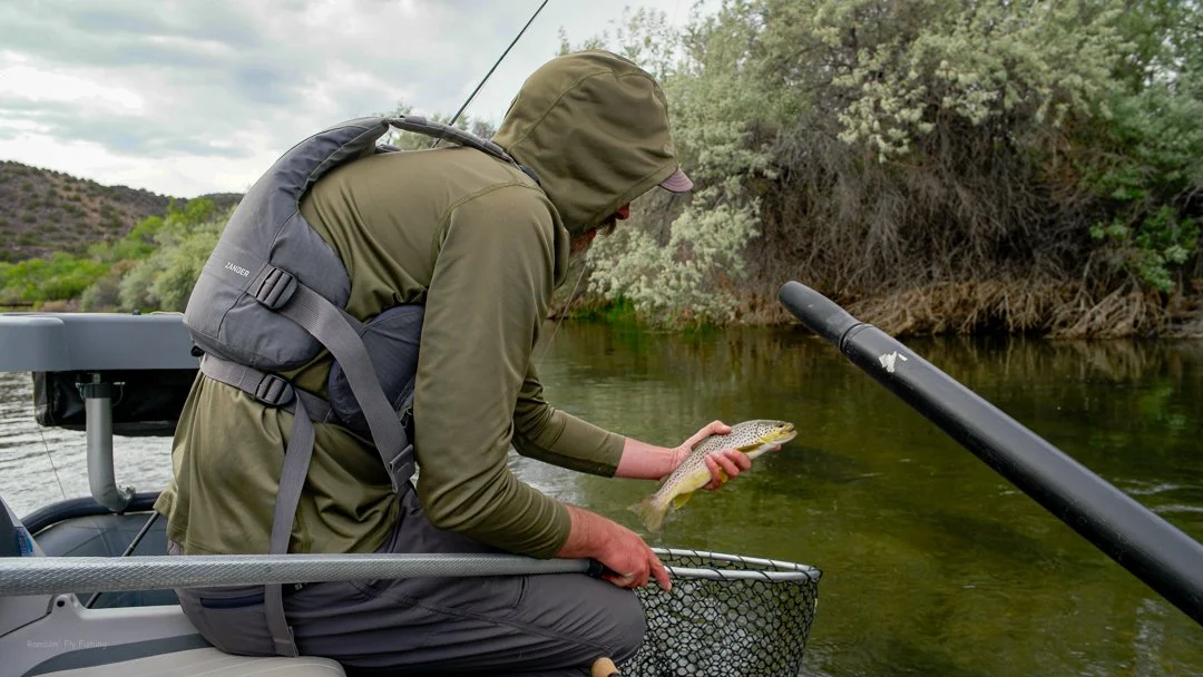 Man in hooded jacket and backpack kneeling in a boat, holding a freshly caught fish over a net, near calm river with trees and bushes in the background.