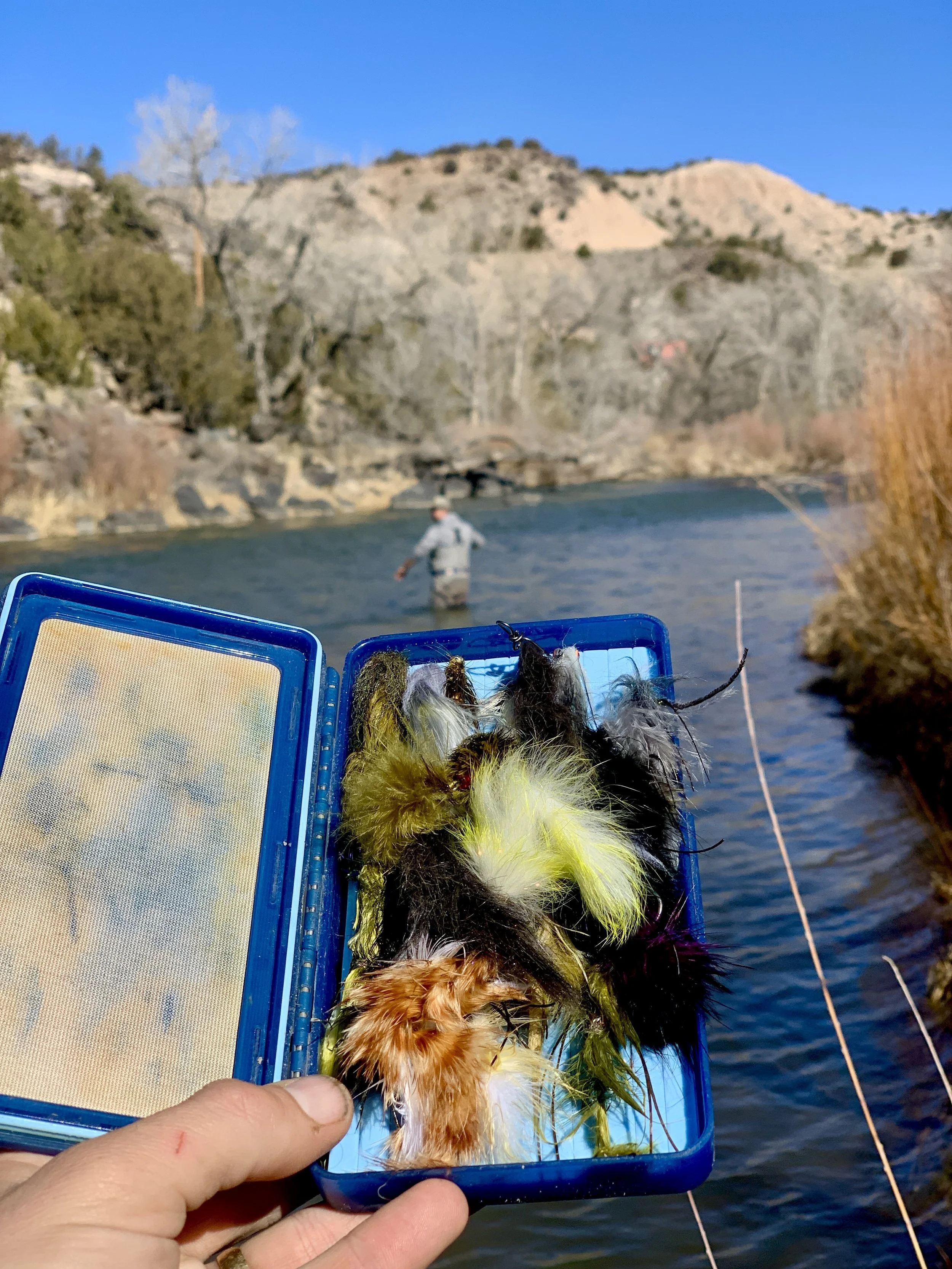A person holding a tackle box with colorful fishing lures in front of a river, with a person fishing in the water and a mountainous landscape in the background.