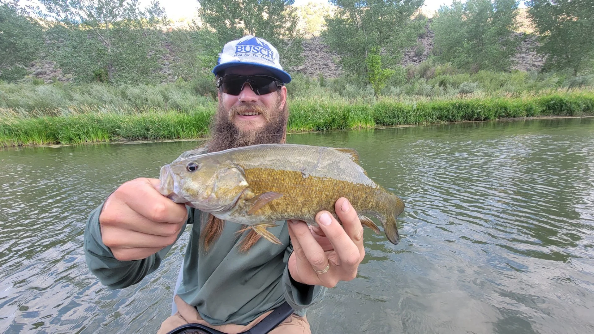 Man with long hair, beard, sunglasses, and a cap holding a large fish in a river with green trees and grass in the background.