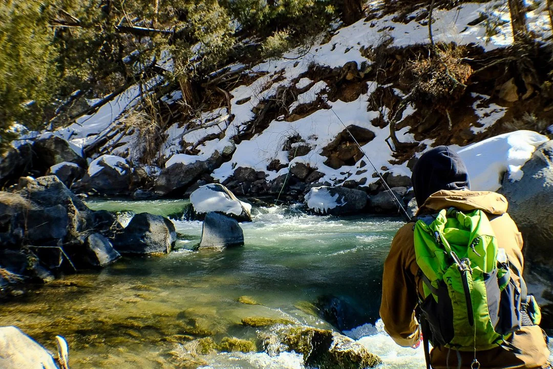 A person fishing in a river with snow on the rocks and trees in the background.