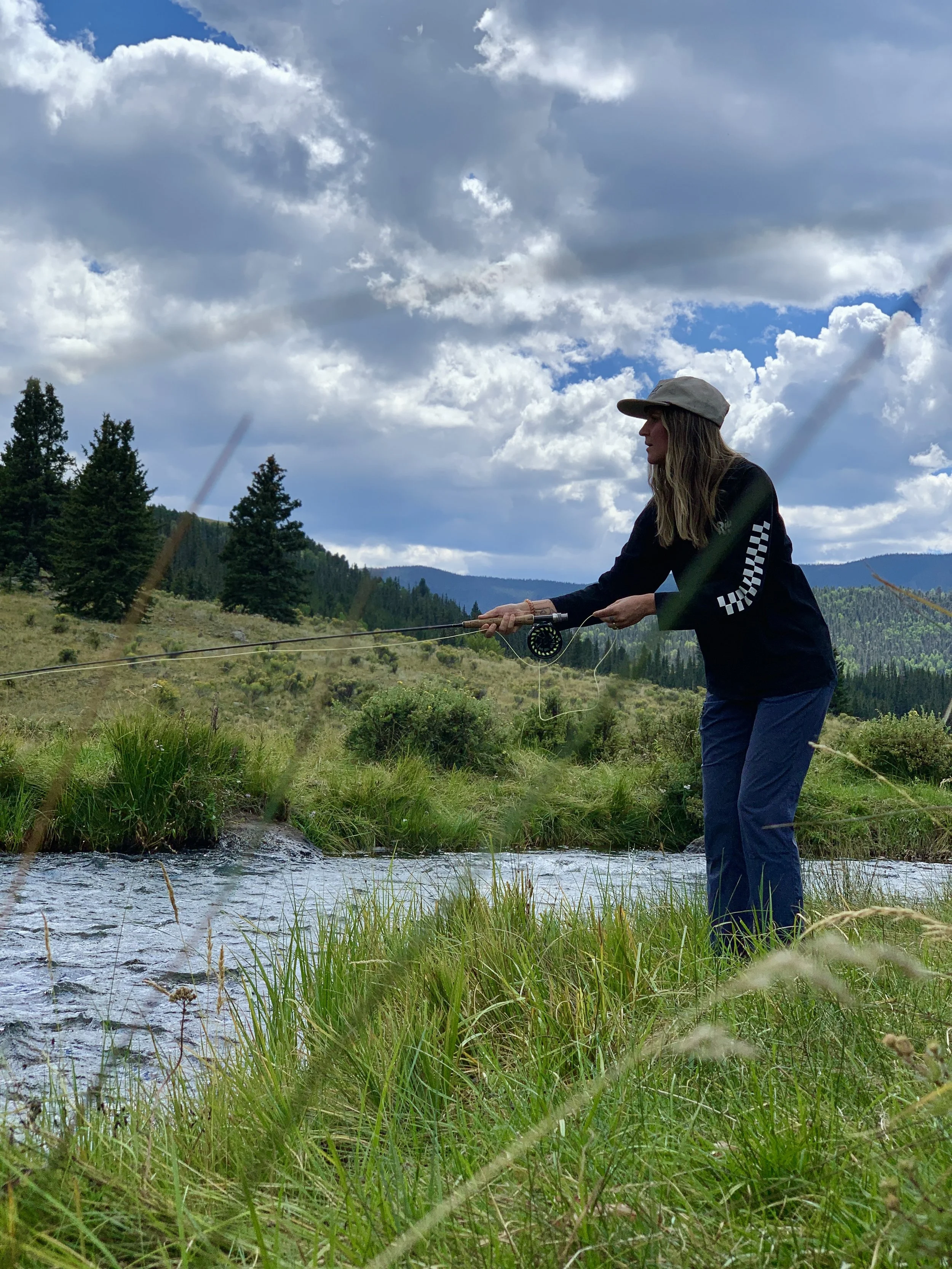 A woman fly fishing in a stream in a mountainous landscape with tall pine trees, green grass, and a cloudy sky.