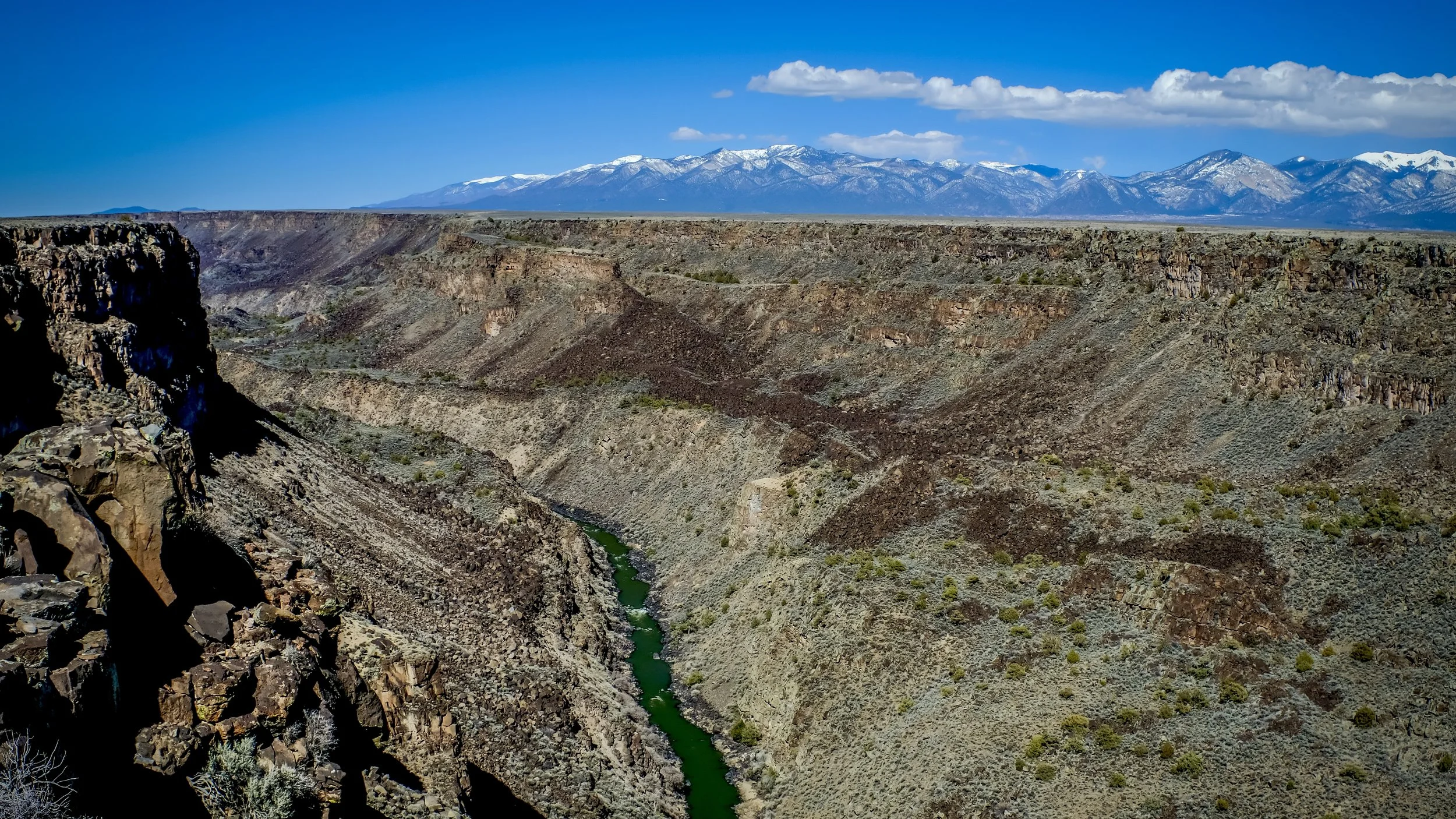 A deep canyon with rugged rocky walls and sparse desert vegetation, with a narrow green river at the bottom, and snow-capped mountains in the background under a bright blue sky with scattered clouds.
