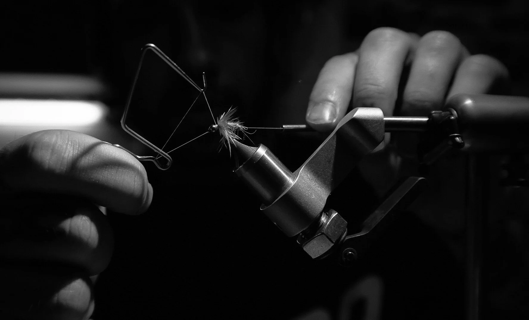 A person using a fly tying vise to craft a fishing fly, with focus on their hands and the fishing hook with feathers.