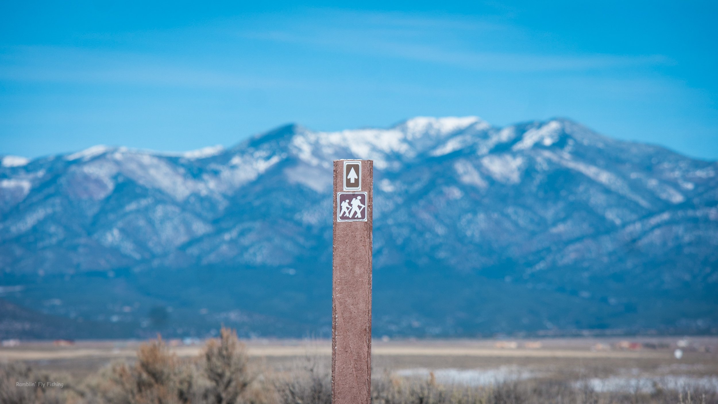 A trail marker pole with an arrow pointing upward and icons of hikers, against a backdrop of snow-capped mountains under a blue sky.
