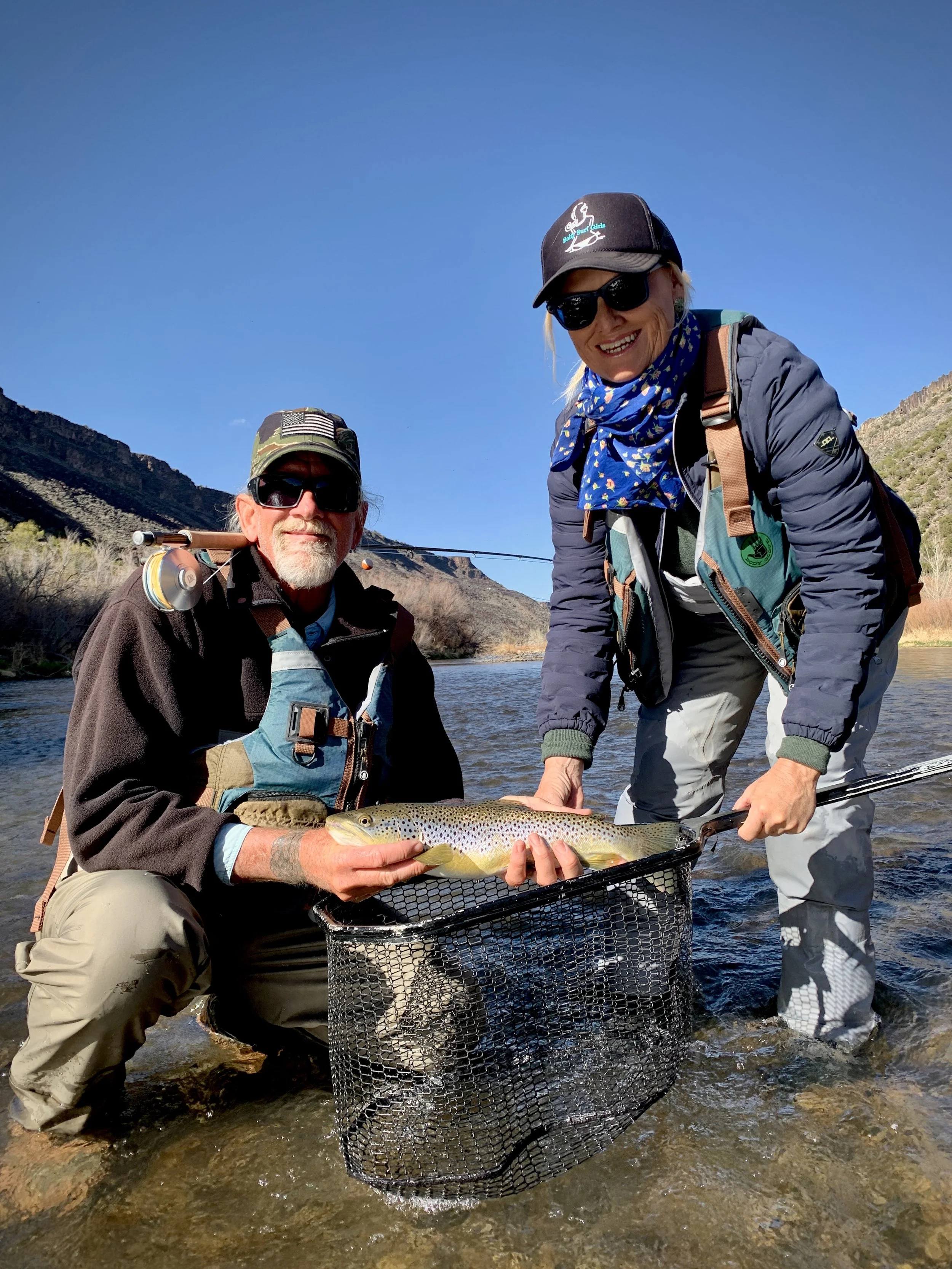 Two people fishing in a stream, holding a large fish over a fishing net, with mountains and a clear blue sky in the background.