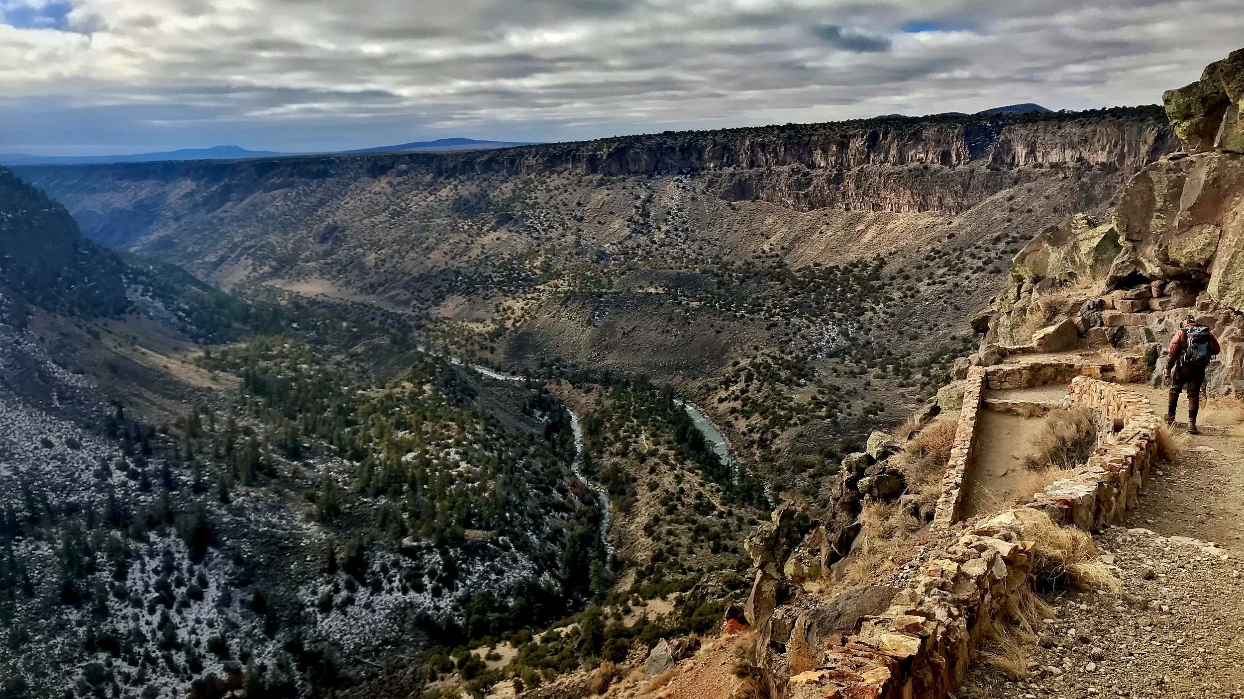 Hiker walking along a rugged trail on a mountain ledge overlooking a deep canyon with a river at the bottom, surrounded by desert shrubs and sparse vegetation under a cloudy sky.