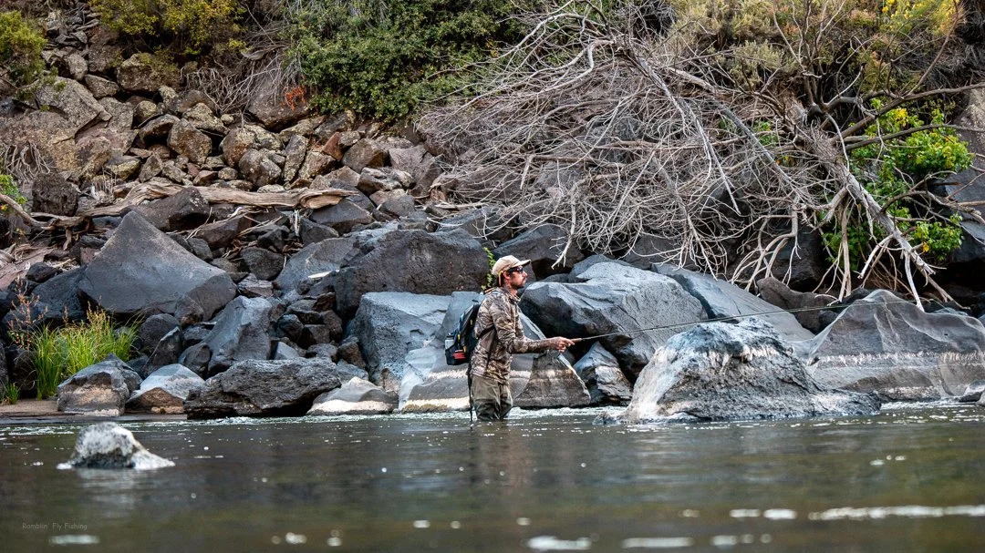 A person standing in a river, fly fishing near large rocks and a rocky, tree-lined bank.